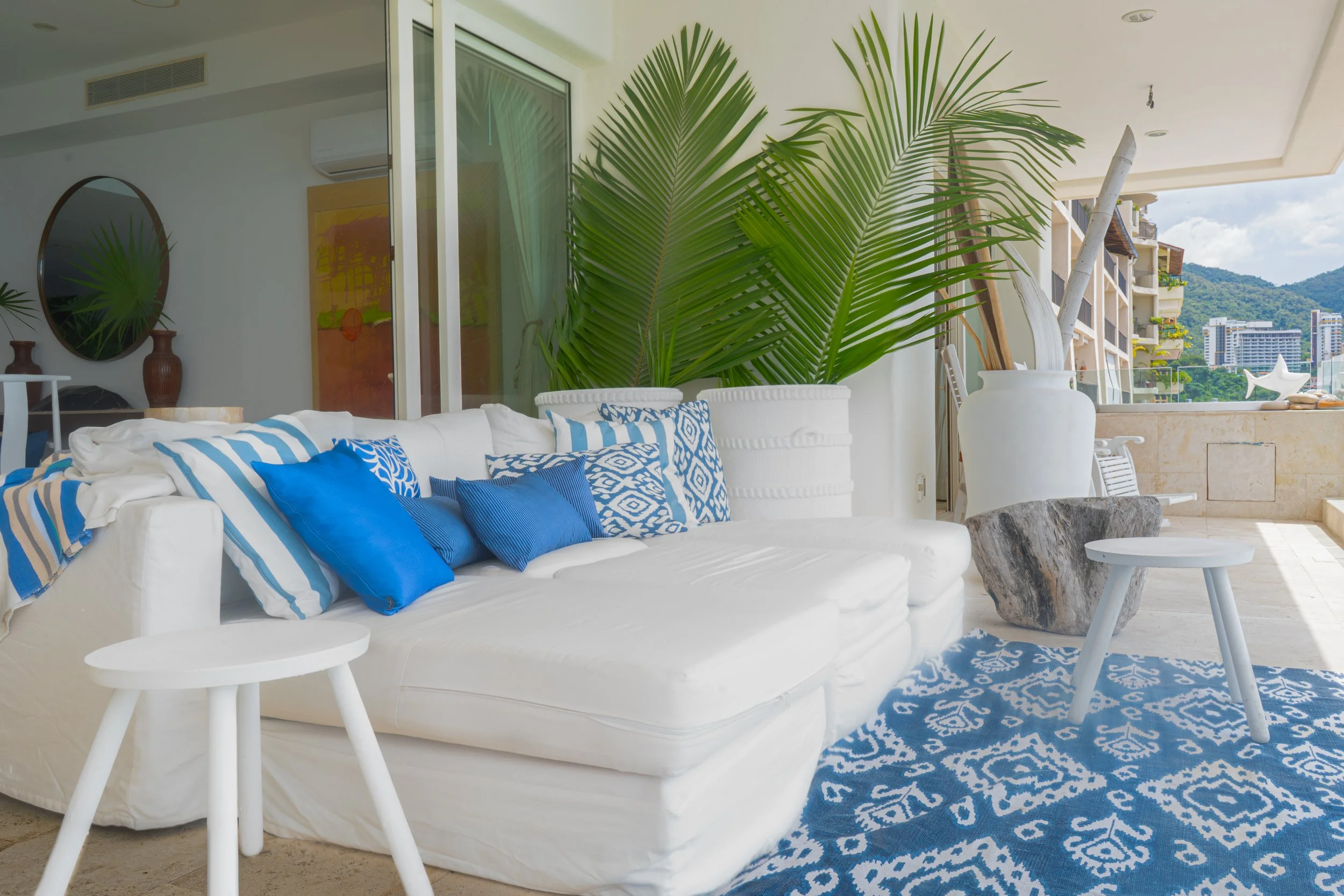 Sala moderna con sofá blanco, cojines azules y decoraciones tropicales, con vistas a la ciudad y montañas al fondo.