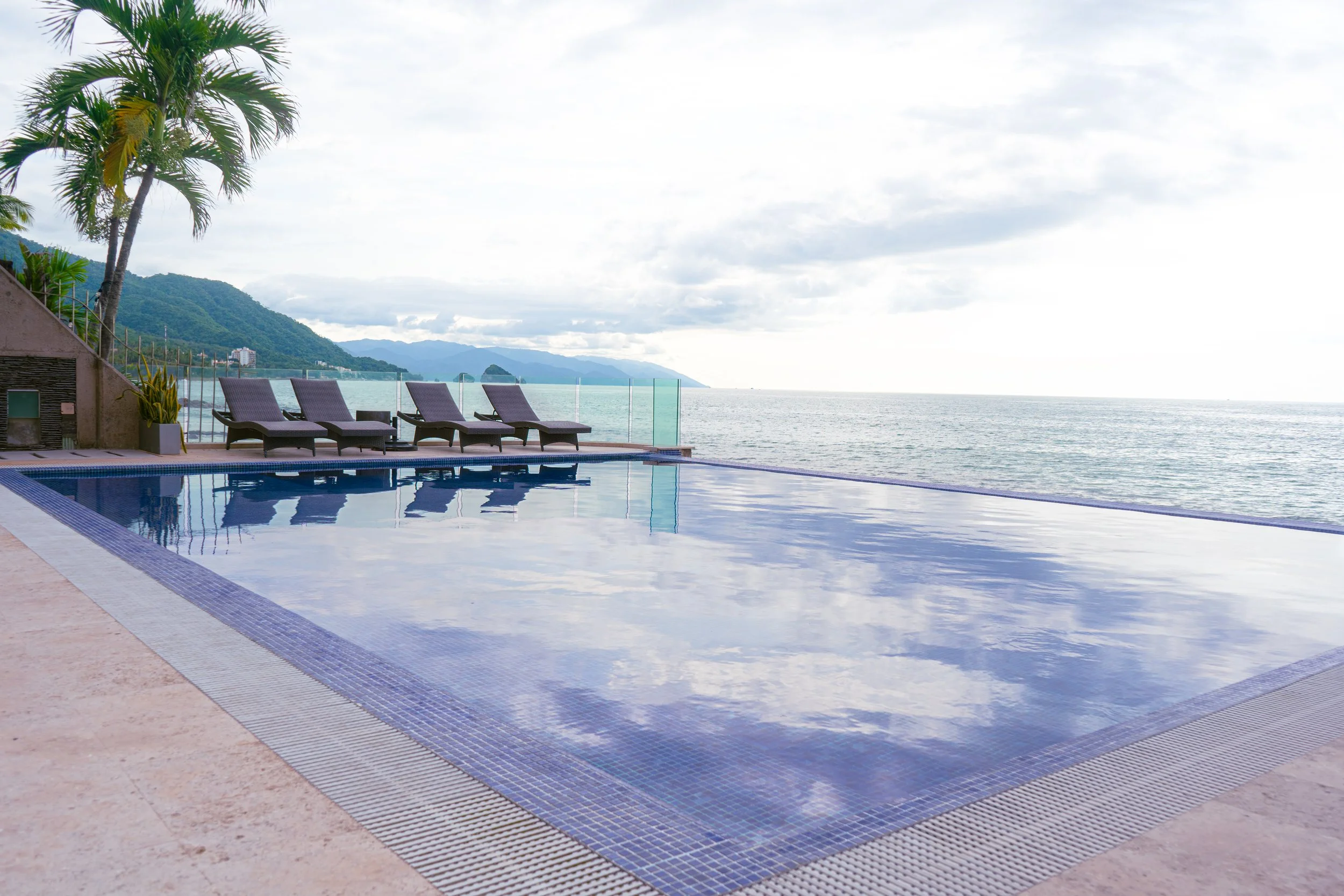 Piscina en un hotel con vista al mar, palmeras y tumbonas en una terraza junto a la piscina