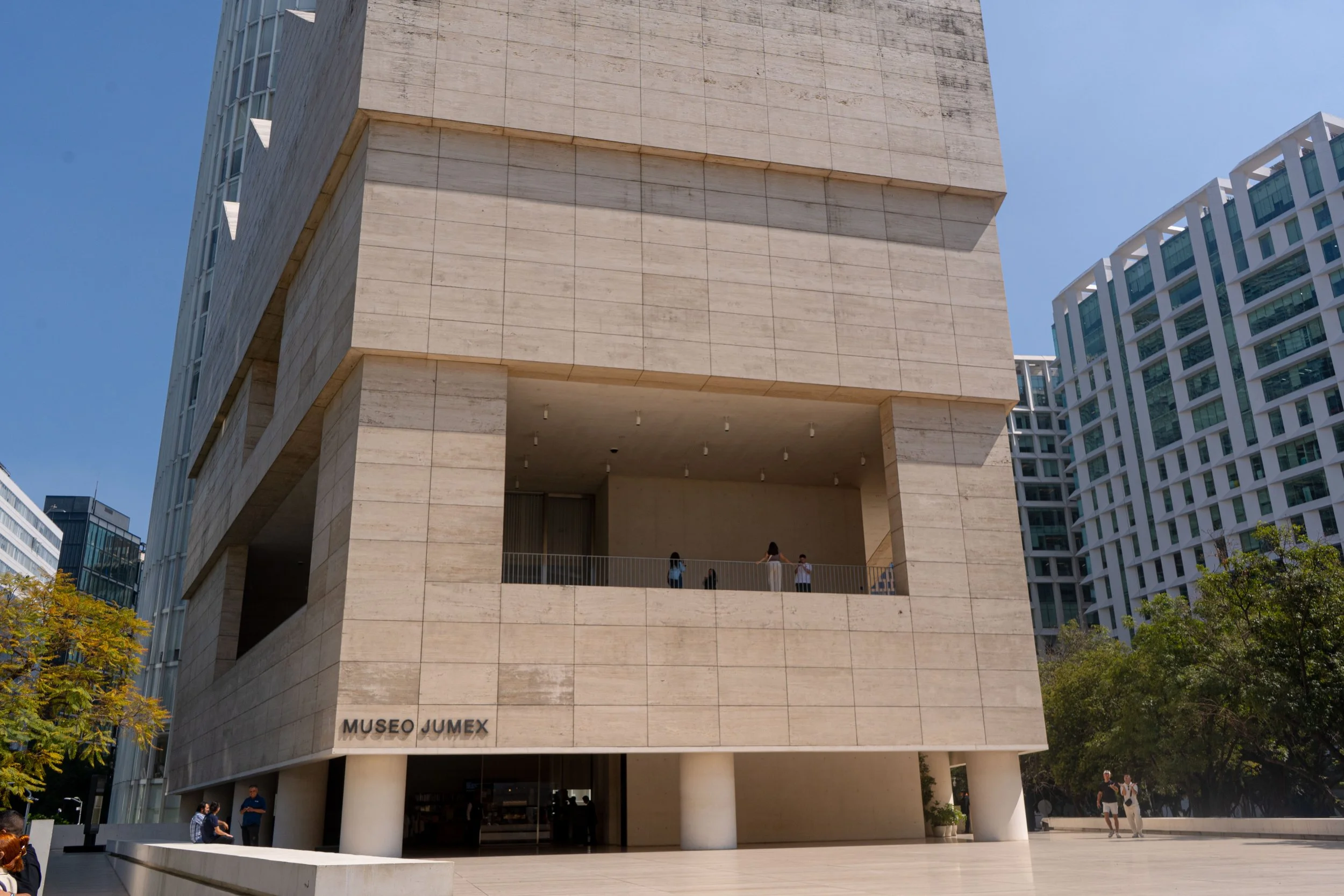 Fachada del Museo Jumex en una ciudad moderna con varios edificios altos y árboles. La estructura es de color beige y tiene un diseño contemporáneo con un balcón en la parte superior donde hay varias personas.
