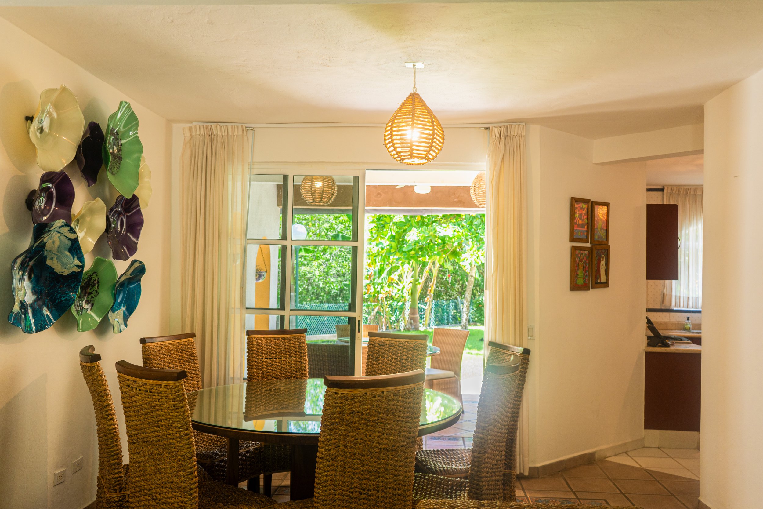 Sala de comedor con mesa de cristal y sillas de mimbre, vista a jardín con árboles verdes, decoraciones en la pared y cortinas claras.