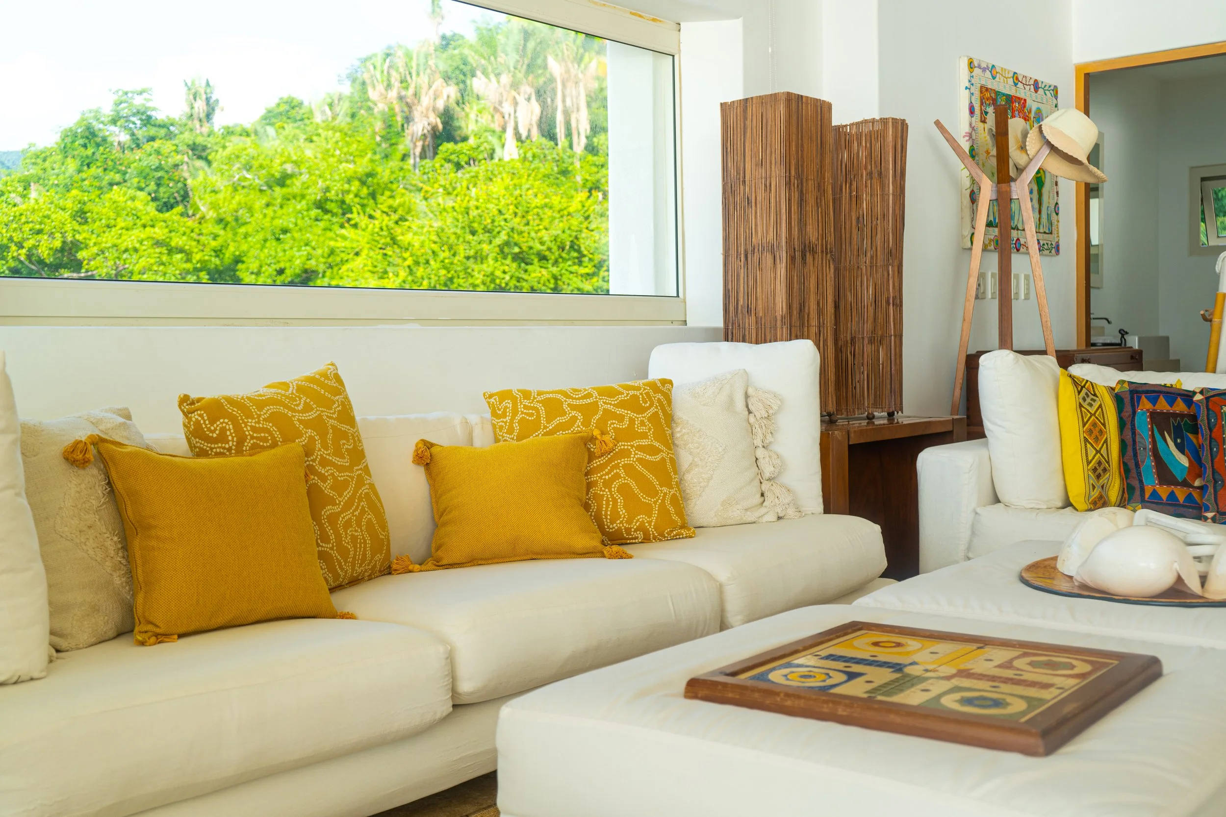 Sala de estar con sillones blancos, cojines amarillos y decoraciones de estilo tropical, ventana grande con vista a árboles verdes.