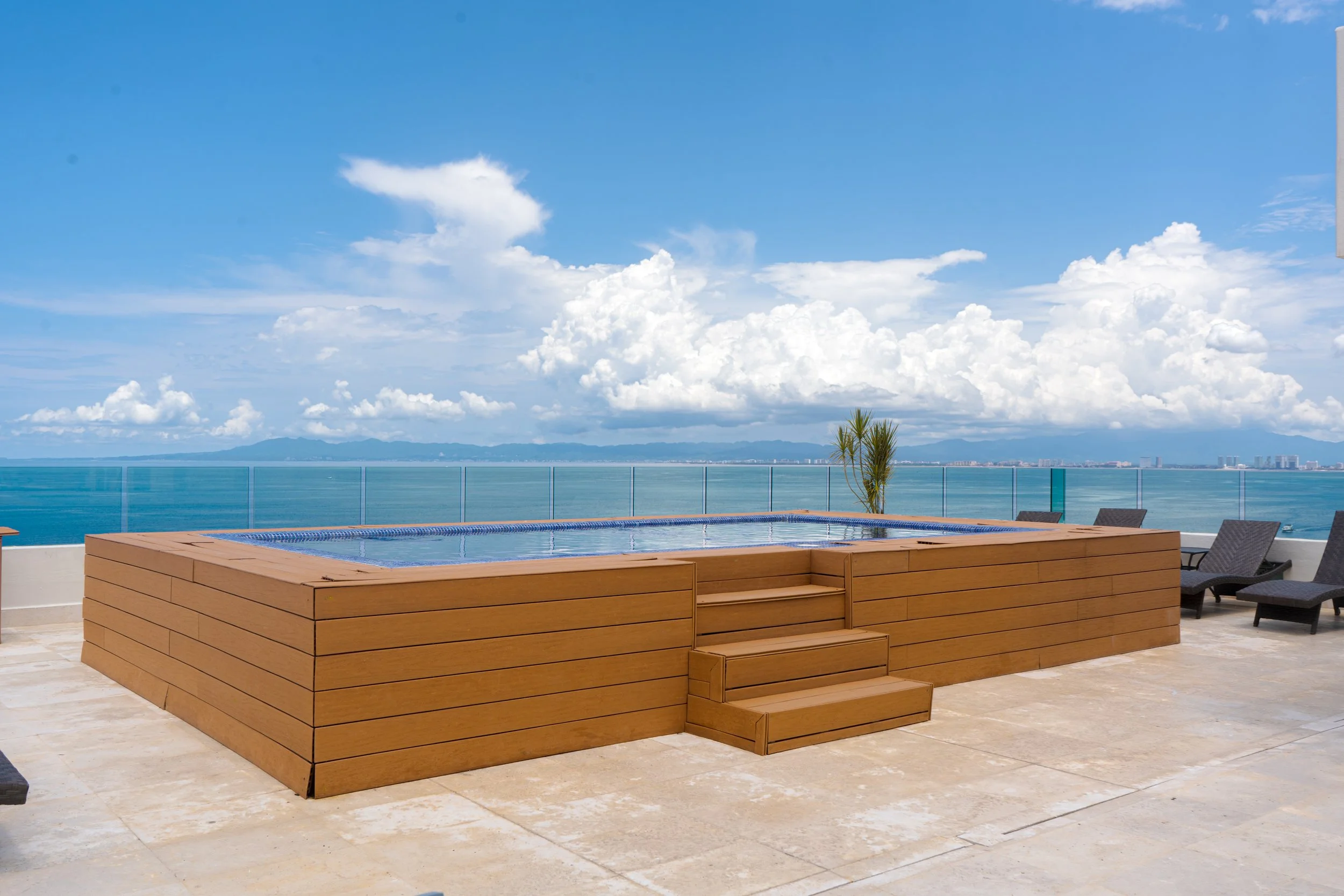 Piscina en una terraza con vista al mar, con silla de descanso y plantas, cielo despejado con nubes.