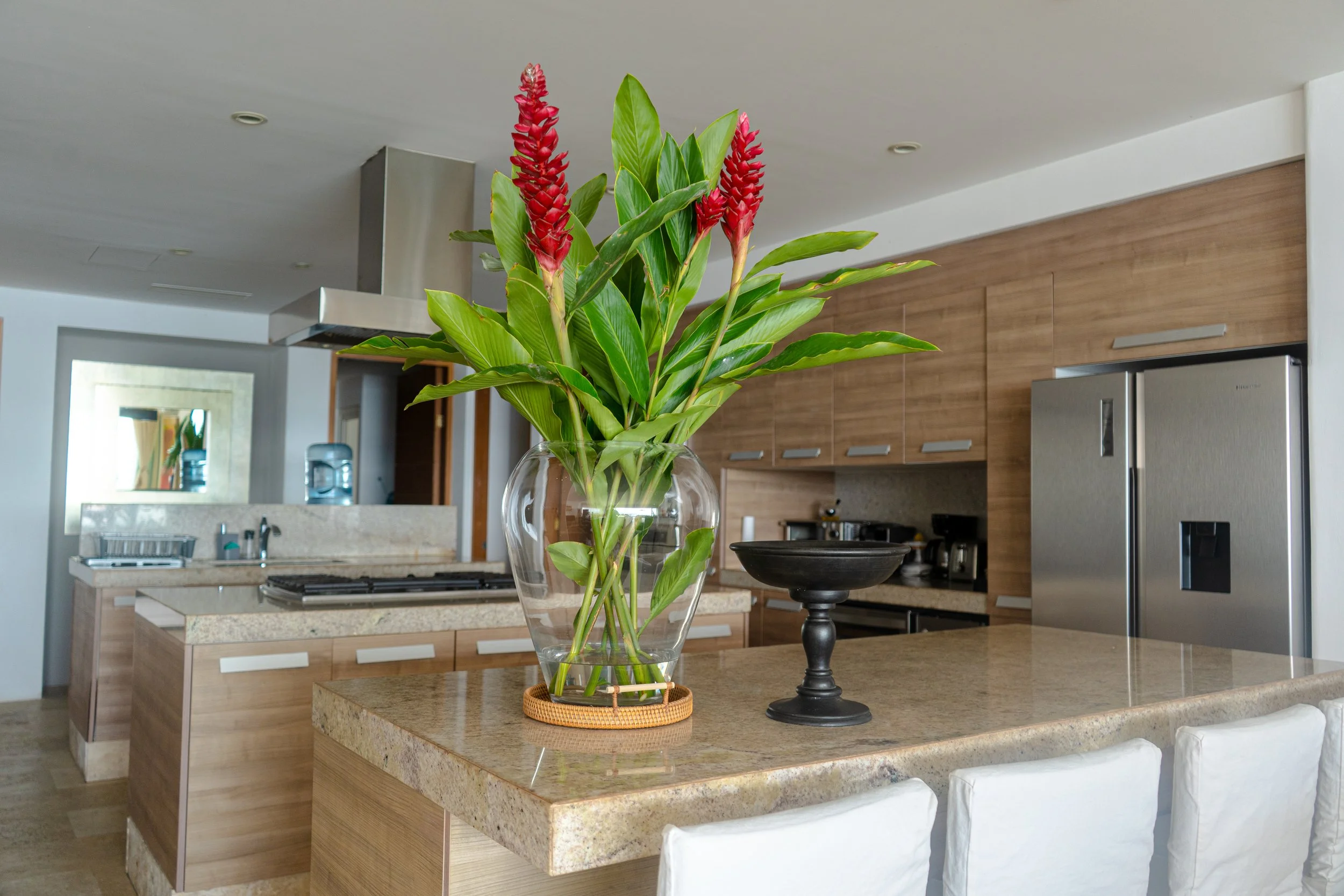 Interior de una cocina moderna con encimeras de granito, gabinetes de madera y electrodomésticos de acero inoxidable, con un florero grande de flores rojas en el centro.