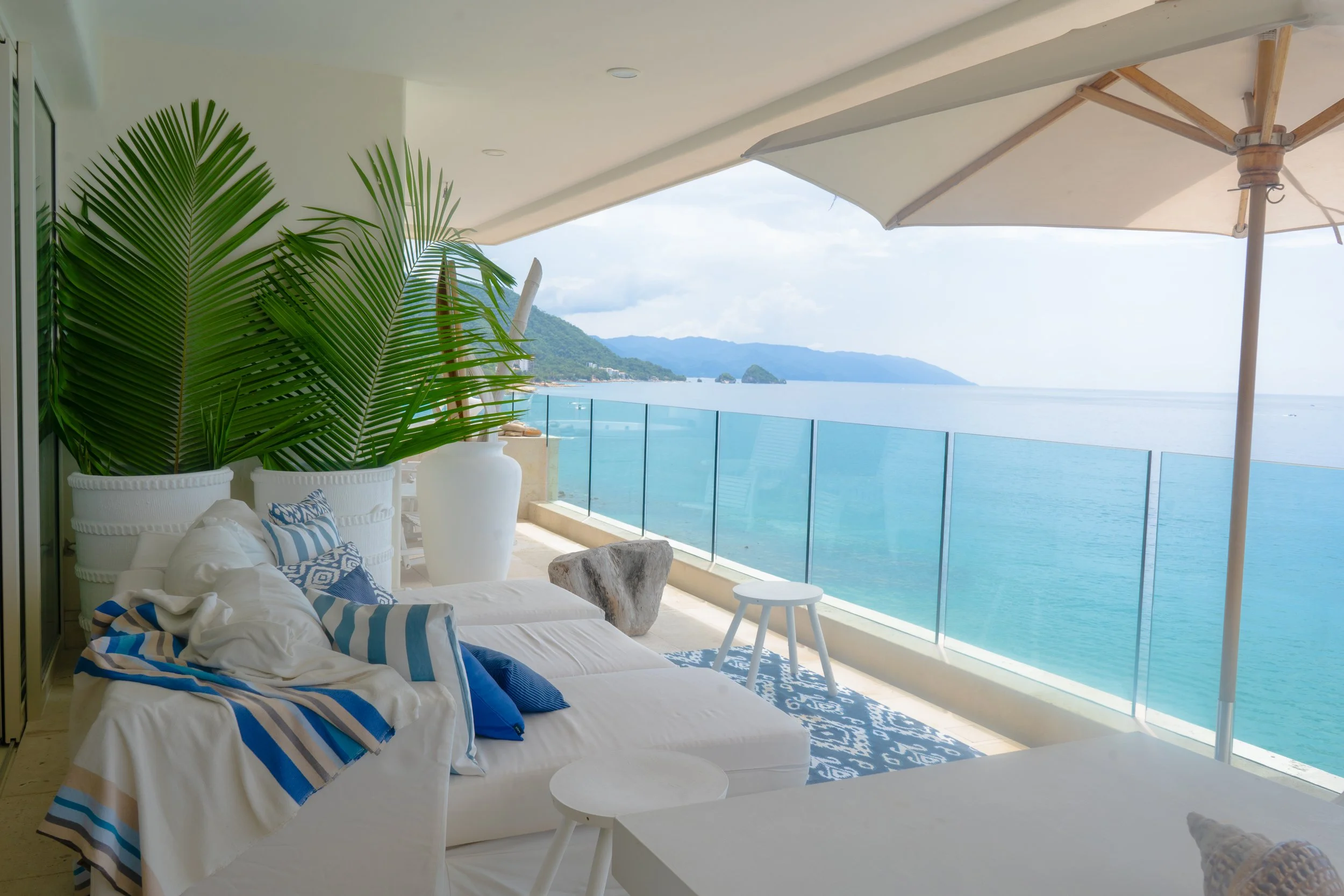 Terraza con vista al mar, área de descanso con sofá y cojines, sombrilla, plantas grandes y decoración tropical.