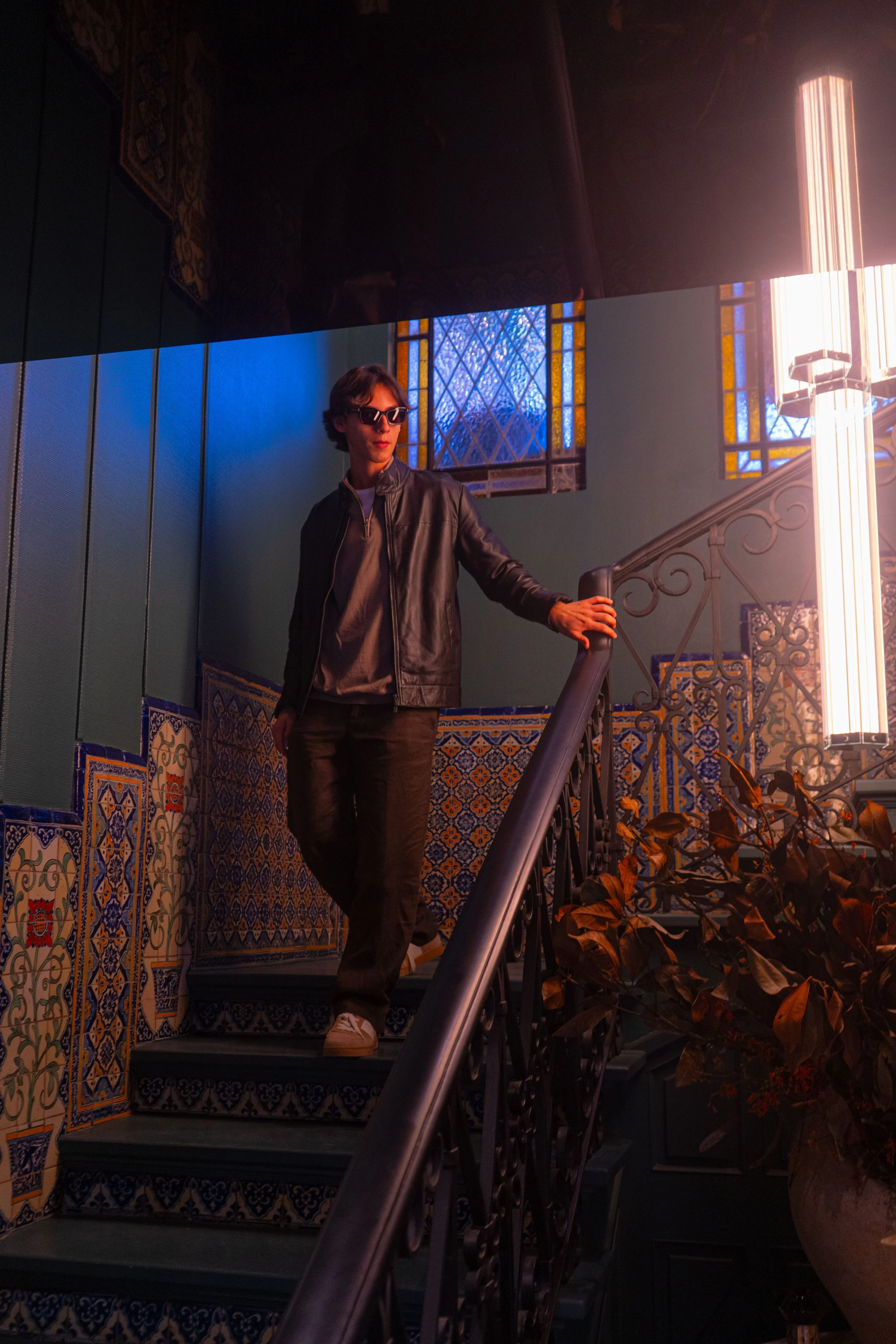 Joven con gafas de sol, chaqueta de cuero, pantalones oscuros y zapatillas, bajando por una escalera decorada con azulejos coloridos en un interior con ventanas de vitrales y luz brillante.