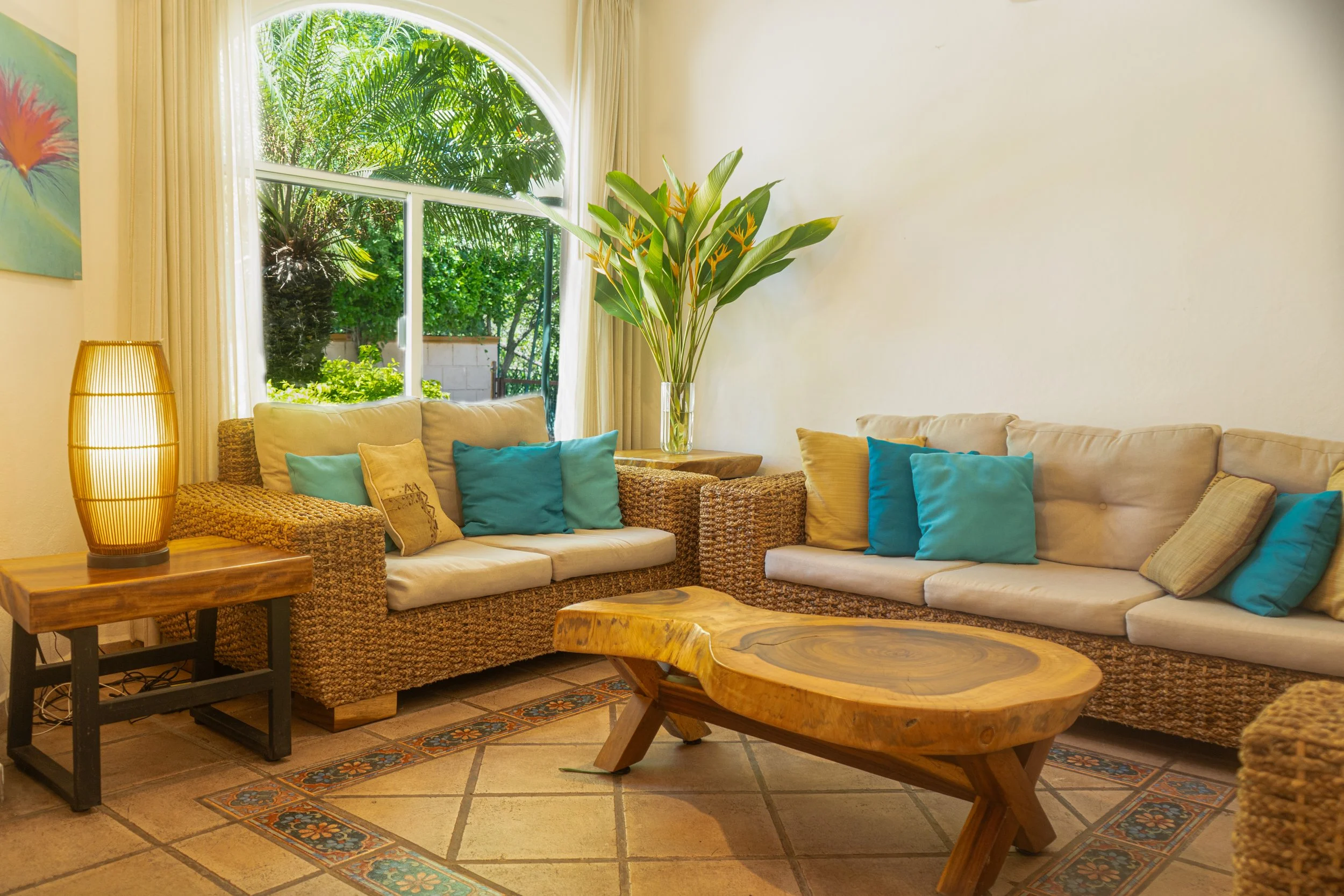 Sala de estar con sofá de mimbre y cojines azules y beige, mesa de madera, lámpara de madera y caña, planta verde y ventana grande con vista a jardín