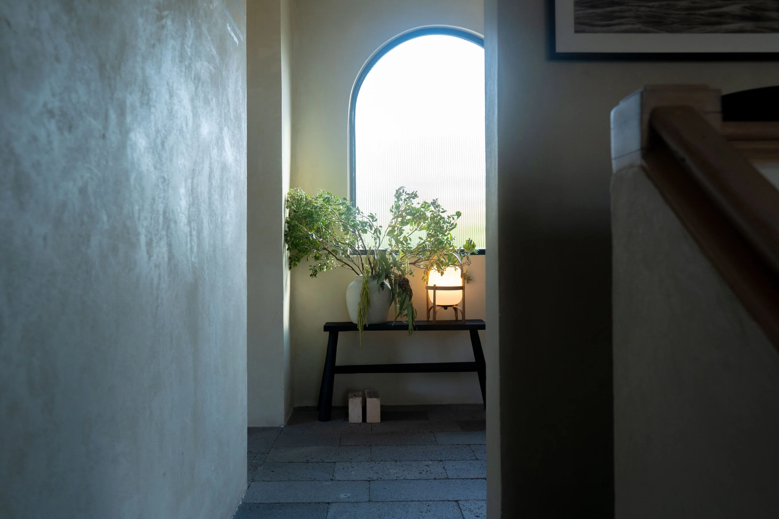 Un pasillo en interiores con paredes beige, piso de baldosas, una ventana arqueada con luz natural y una planta en una mesa negra con lámpara decorativa, en un ambiente tranquilo y minimalista.
