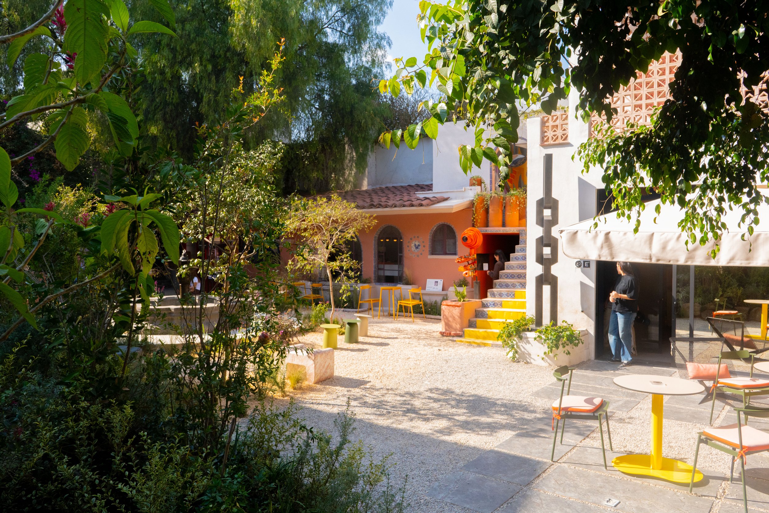 Patio de un café o restaurante con mesas y sillas de colores, rodeado de vegetación y arbustos, con una estructura decorativa color naranja y una mujer de pie en la entrada.