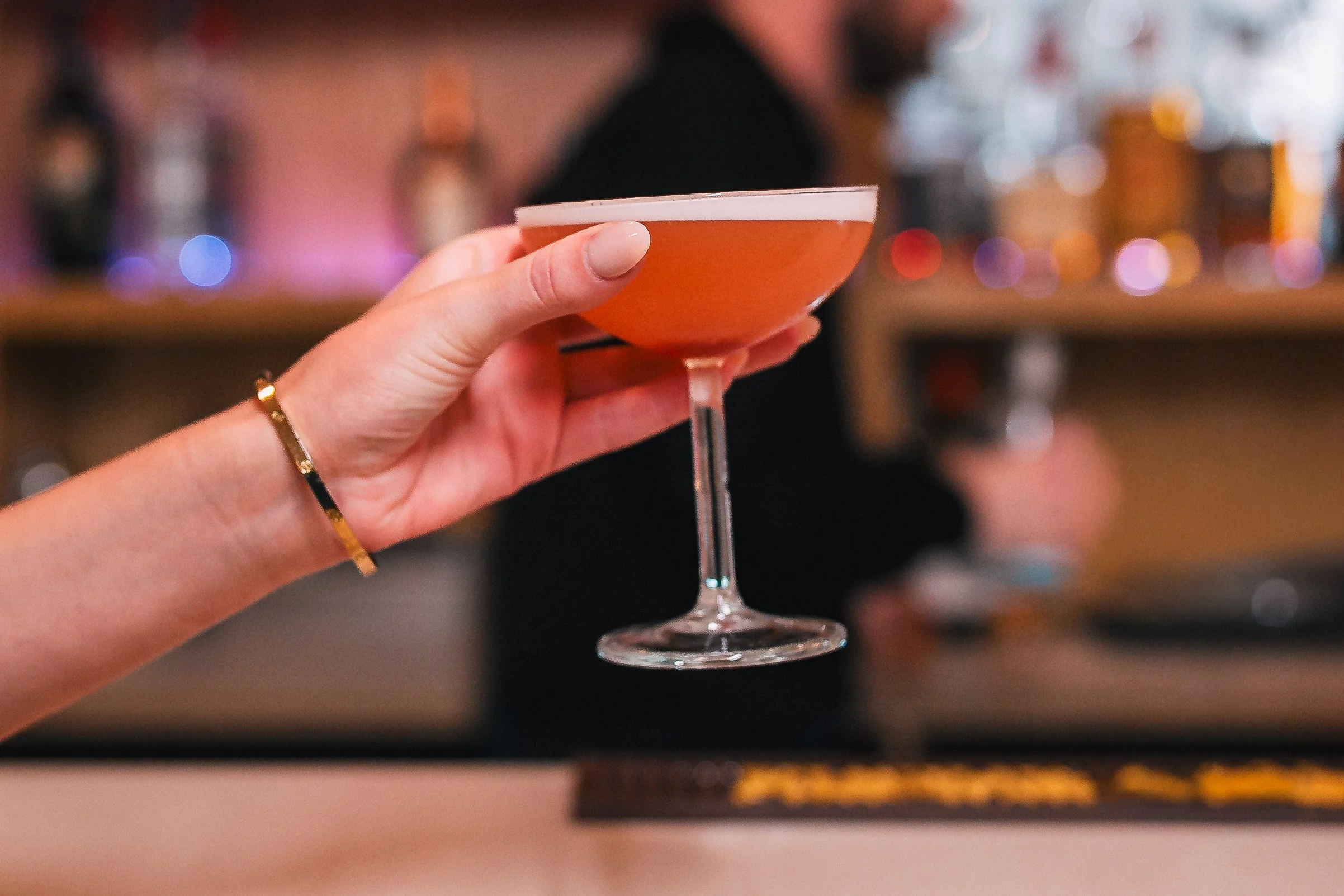 A person holding a pink cocktail in a bayonet glass with a person in the background and colorful bokeh lights.