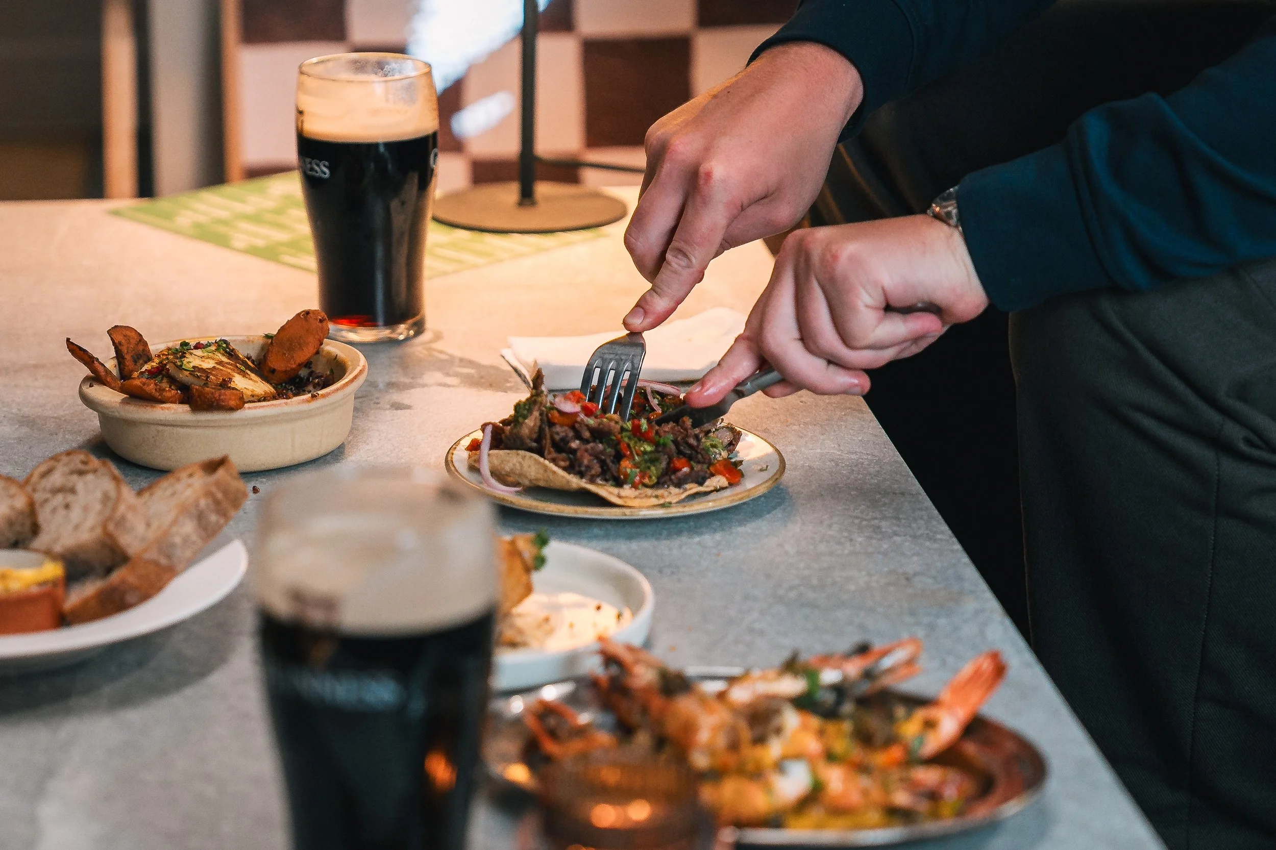 Person serving themselves food from a variety of dishes on a table, including a plate with meat and vegetables, and a glass of dark beer.
