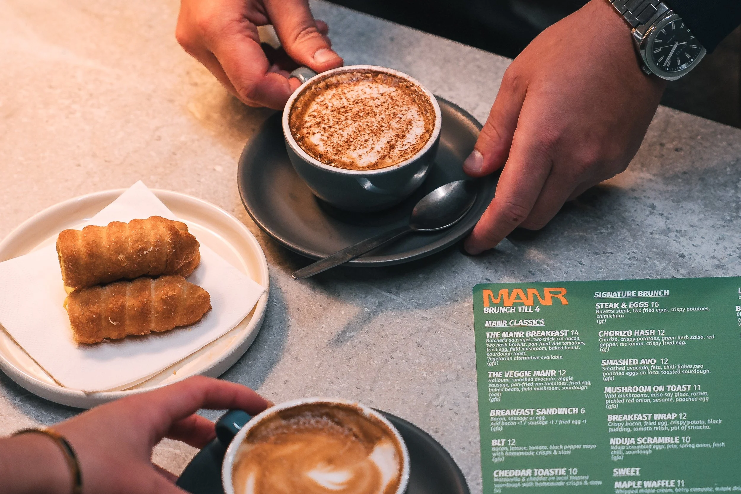 A person is holding a cup of cappuccino topped with foam and cocoa powder, with a spoon on a saucer. On the table, there is a small white plate with two fried spring rolls. Part of a green menu with white text is visible, listing various breakfast and brunch dishes.