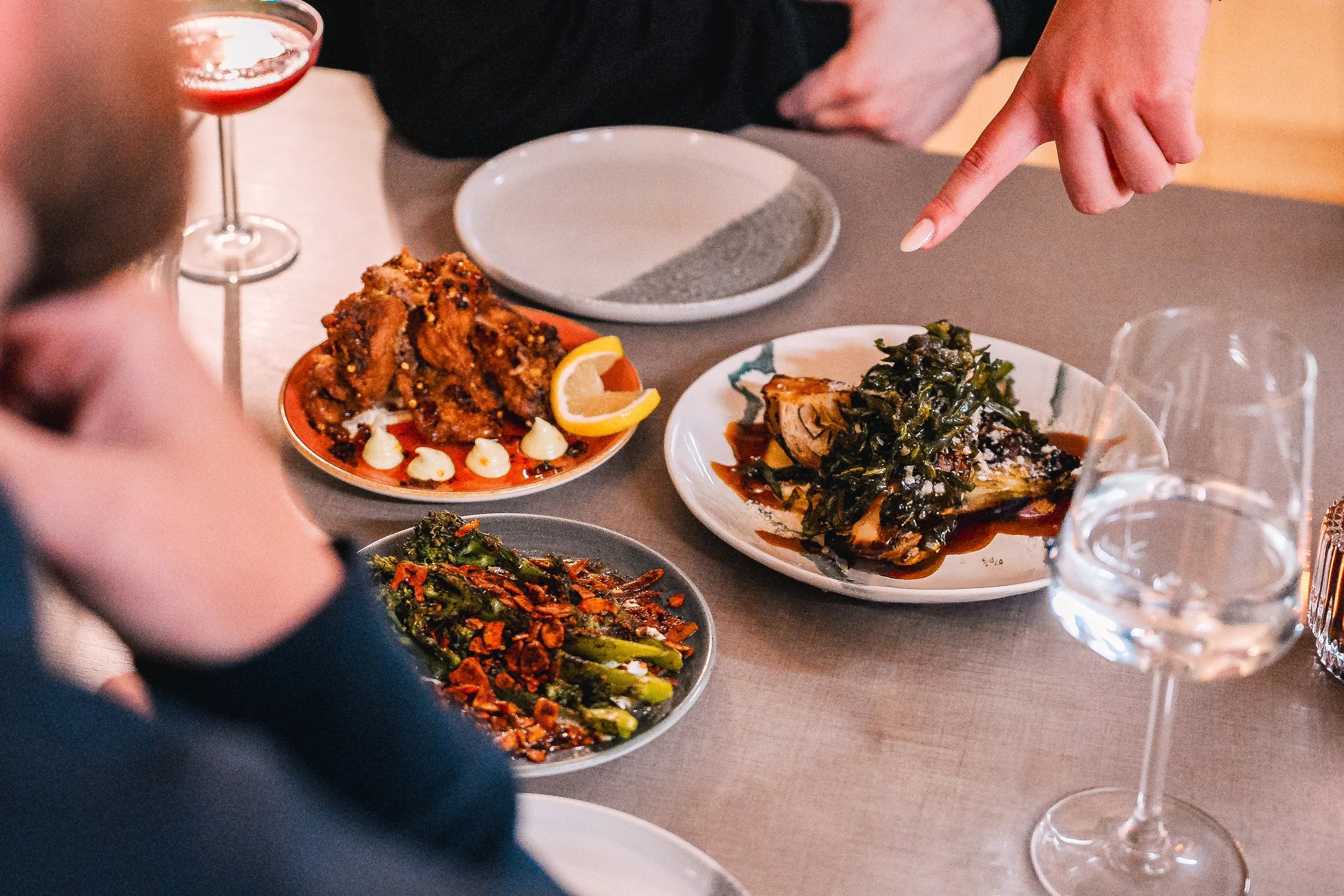 People at a table enjoying a meal with various dishes including fried chicken with lemon wedges, cooked greens, and other vegetables, along with glasses of red wine.