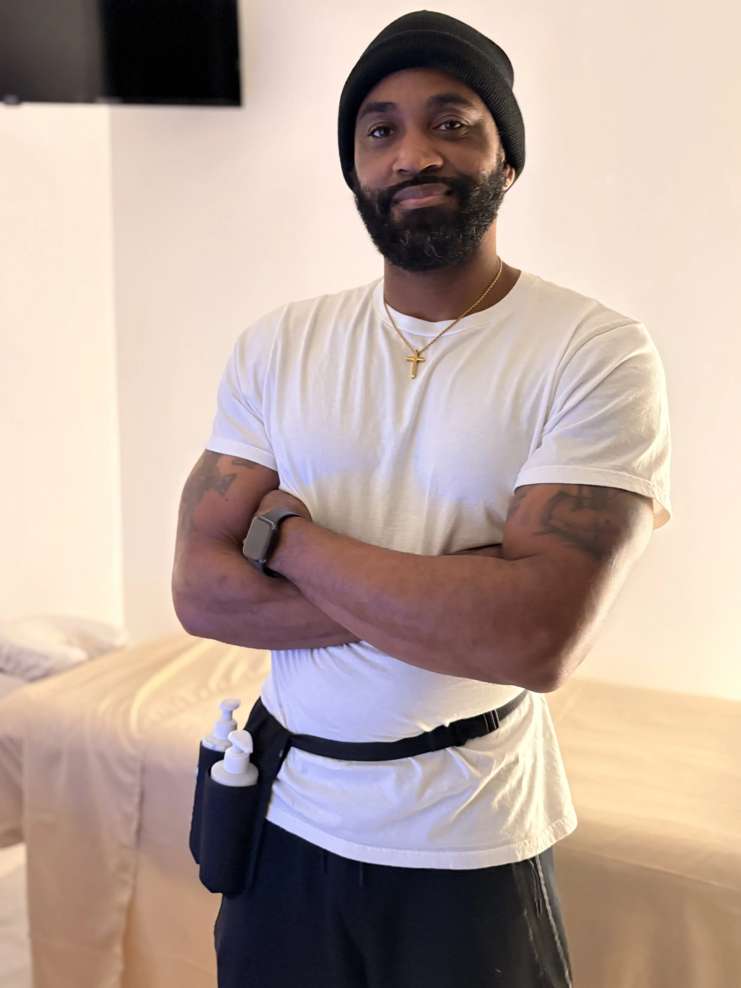 A man with a beard wearing a black beanie, white t-shirt, black pants, and a gold cross necklace, standing with arms crossed in a room with a beige bed and a wall-mounted TV.
