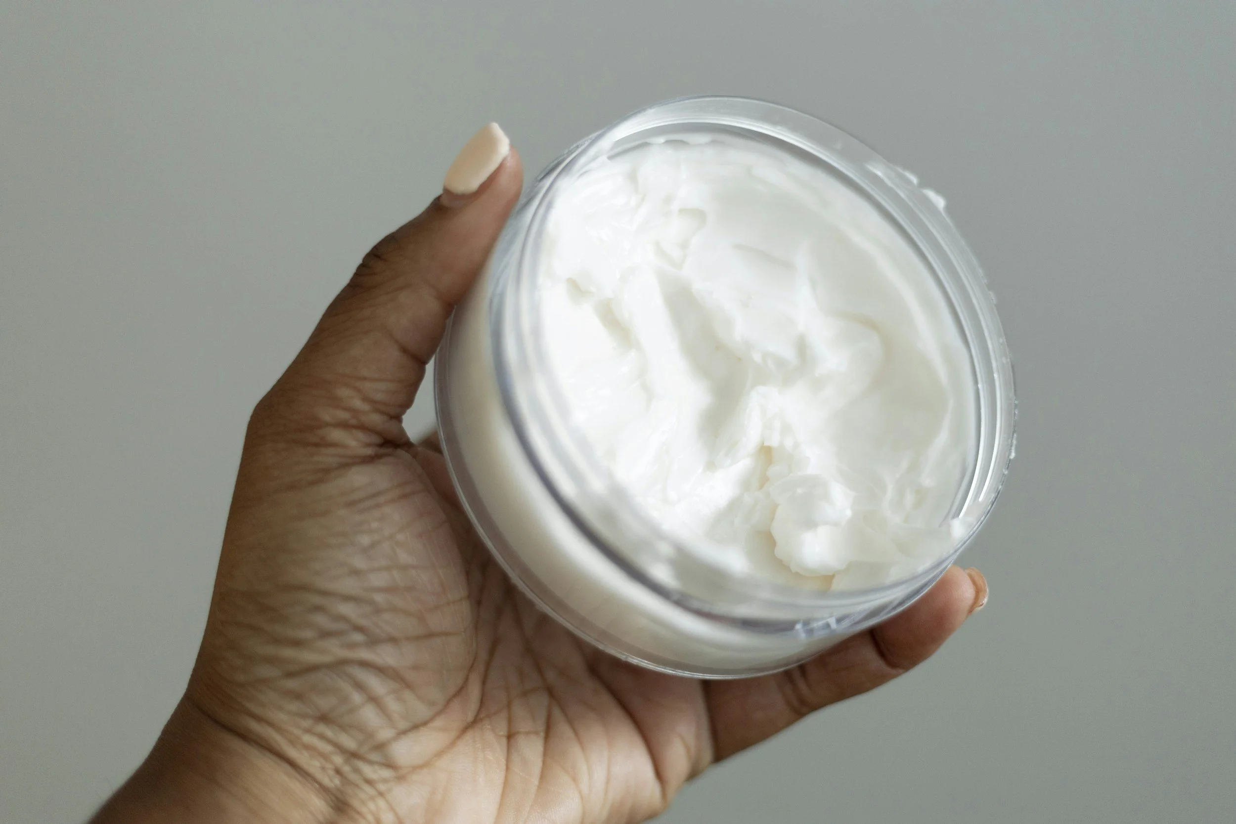 A person holding a clear container filled with white cream or lotion, viewed from above against a plain background.