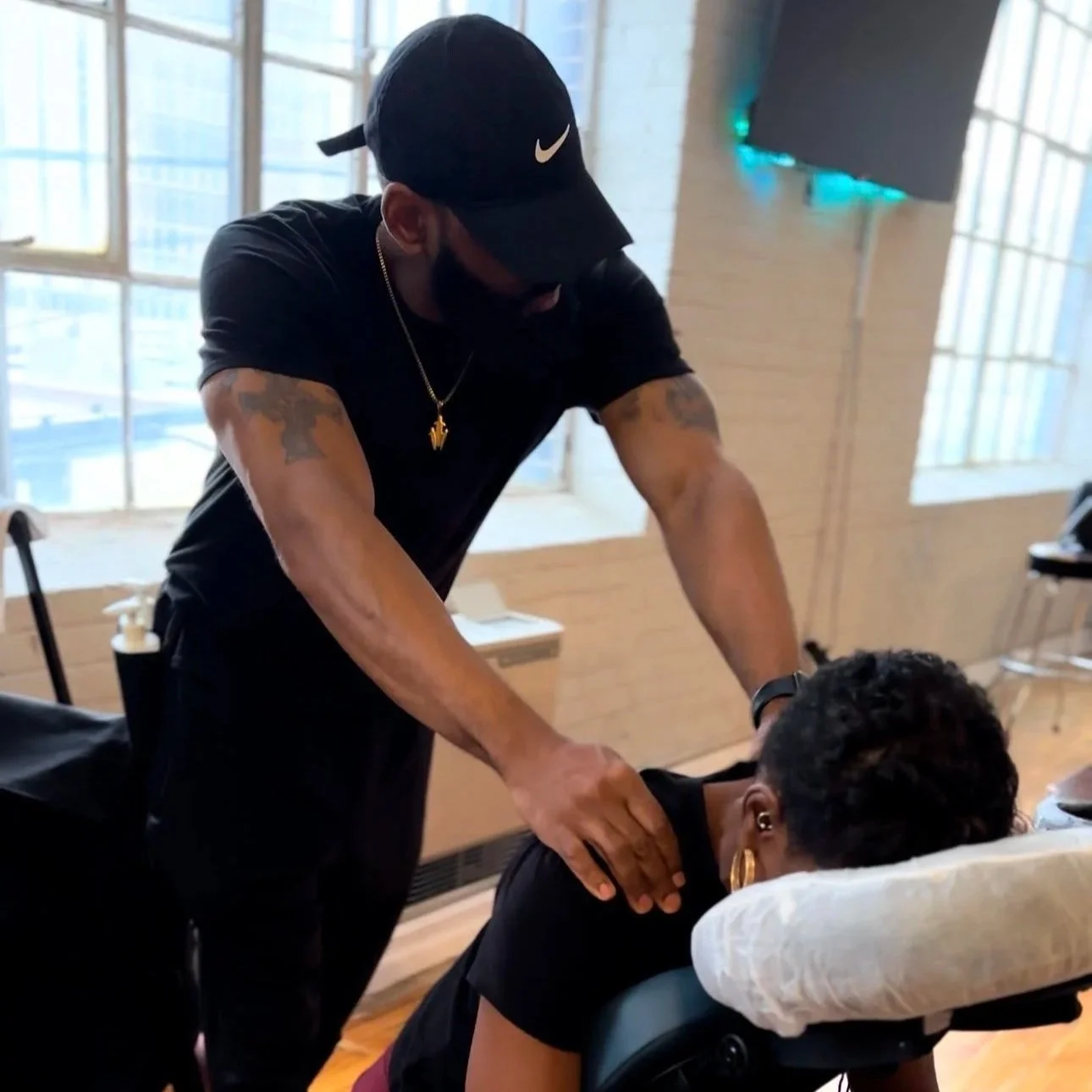 A man providing a massage to a woman laying face down on a massage table in a bright room with large windows.