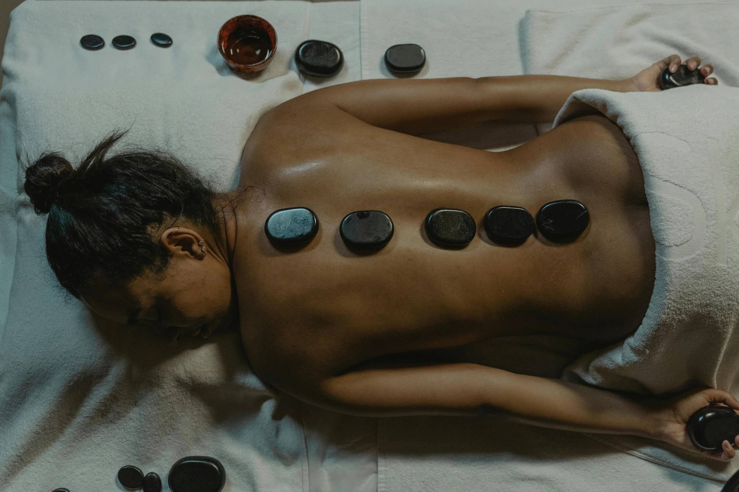 A woman receiving a hot stone massage on her back, lying face down on a massage table with towels, surrounded by massage stones and bowls.