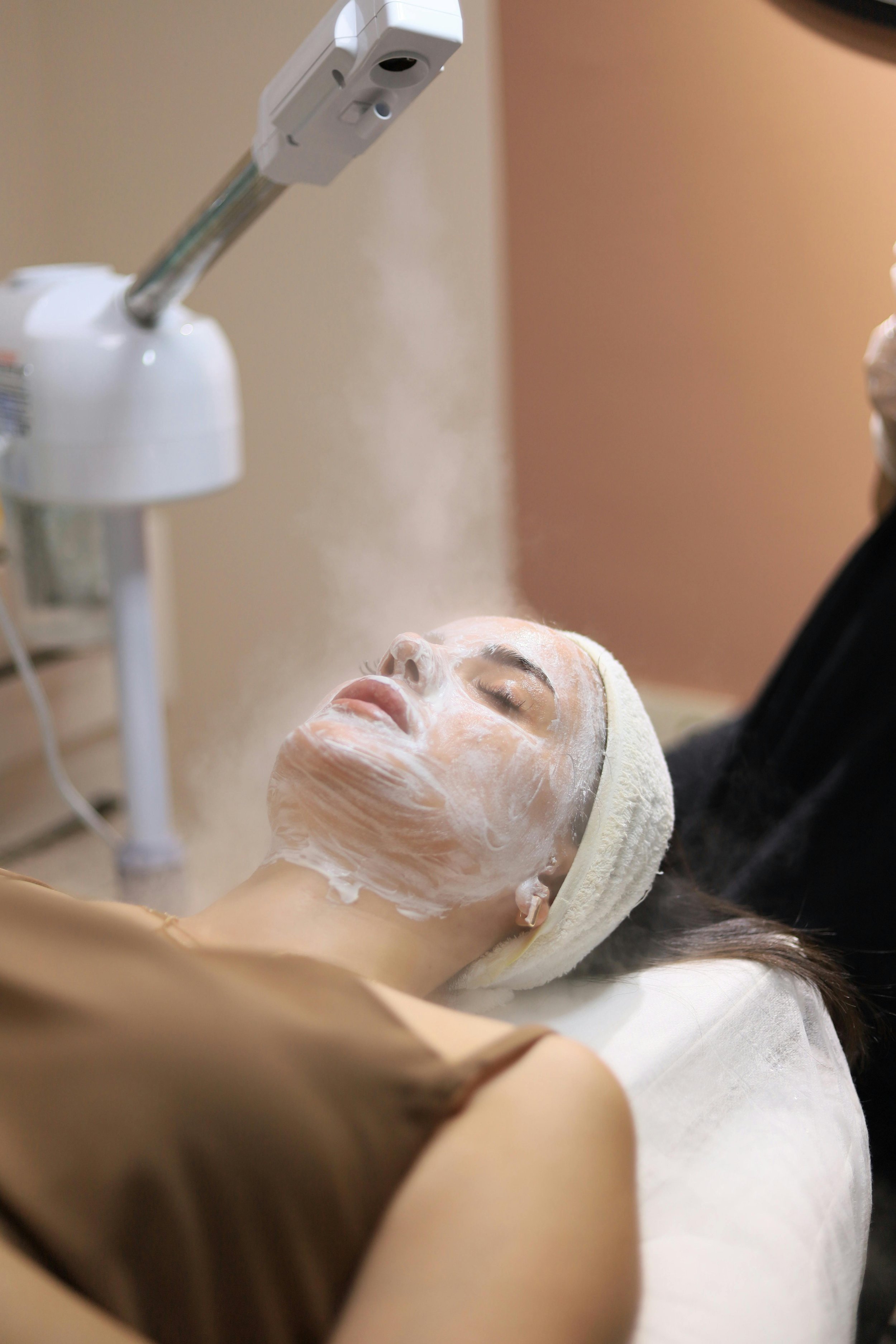 A woman receives a facial treatment with a face mask while lying down with a towel wrapped around her head, and a machine dispensing steam above her face.