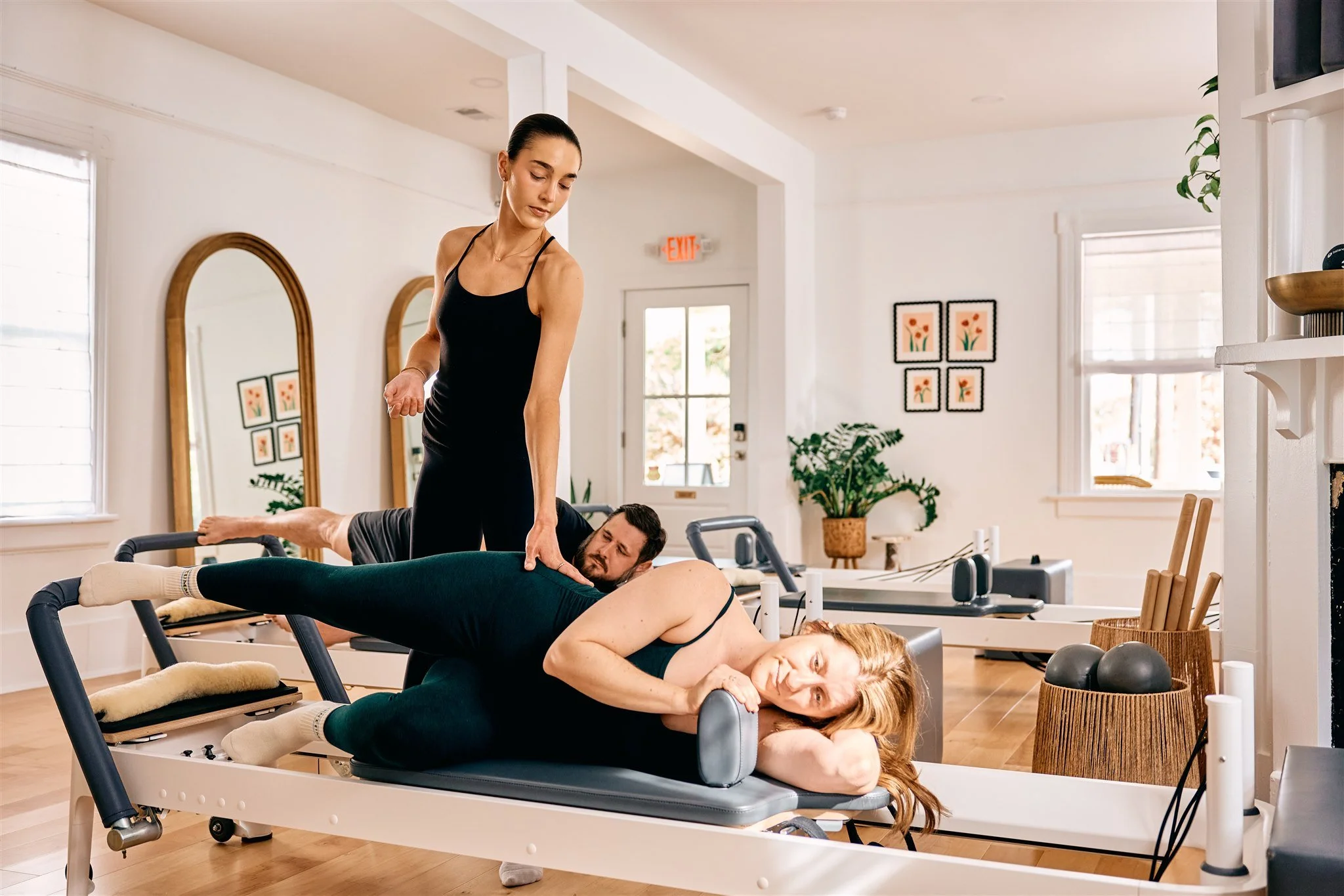 Instructor helping a woman with her form during a pilates exercises in a bright room with mirrors and a fireplace.
