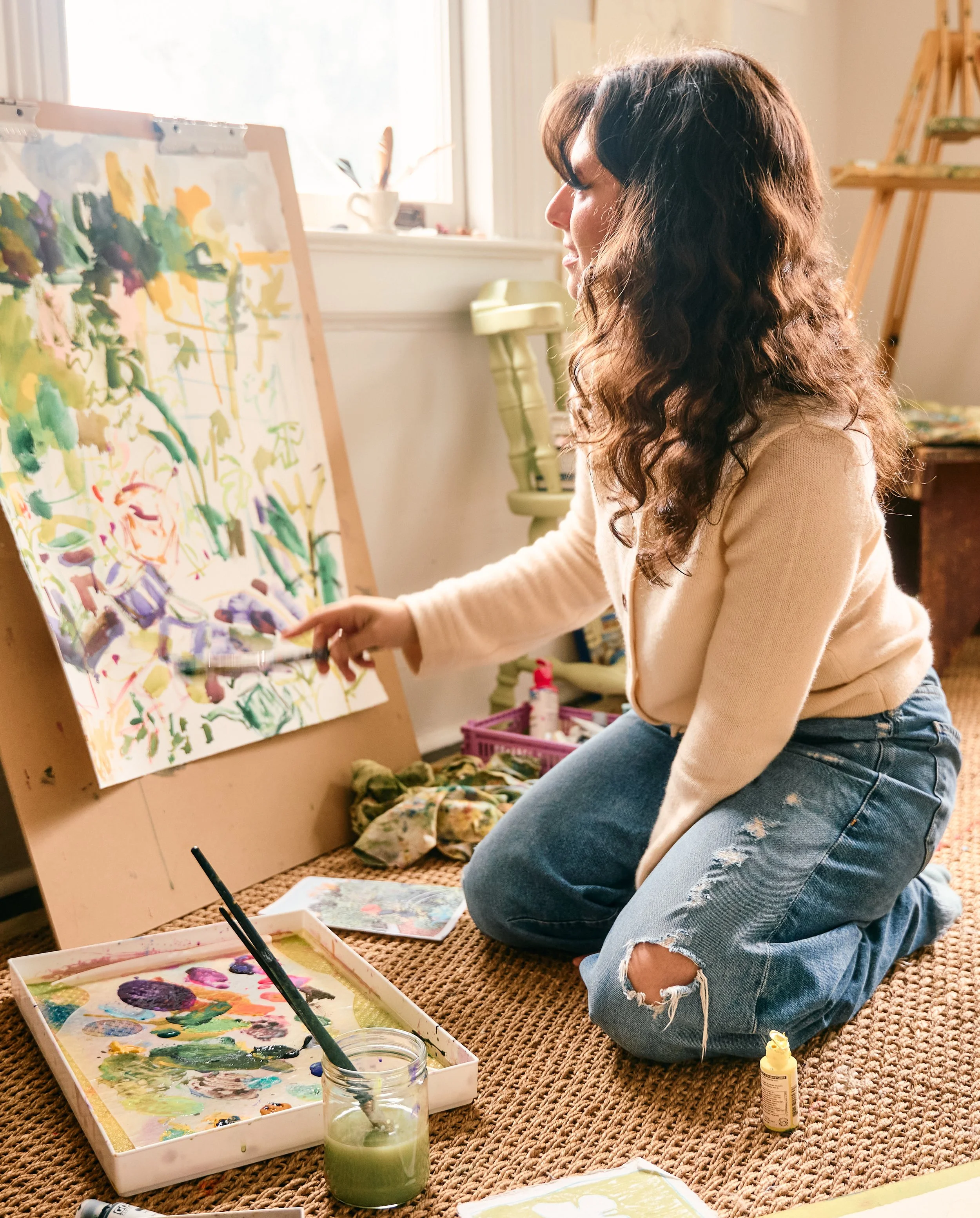 woman kneeling on the floor of a brightly lit room painting a garden scene