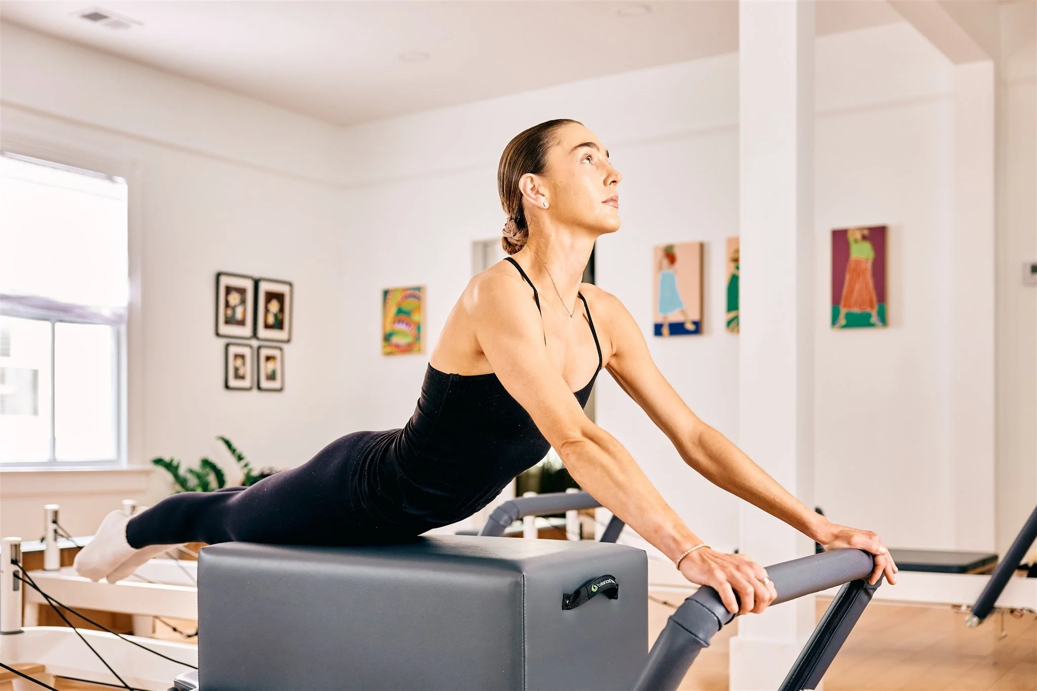 Woman performing swan on a Pilates reformer in a bright room with lots of art work on the walls