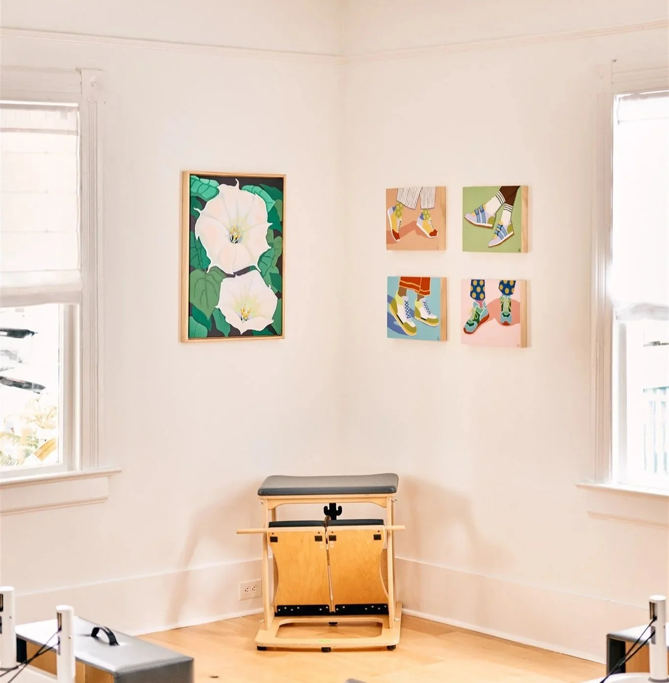 A pilates chair in a corner framed by a painting of flowers, and four bright painting of colorful shoes.