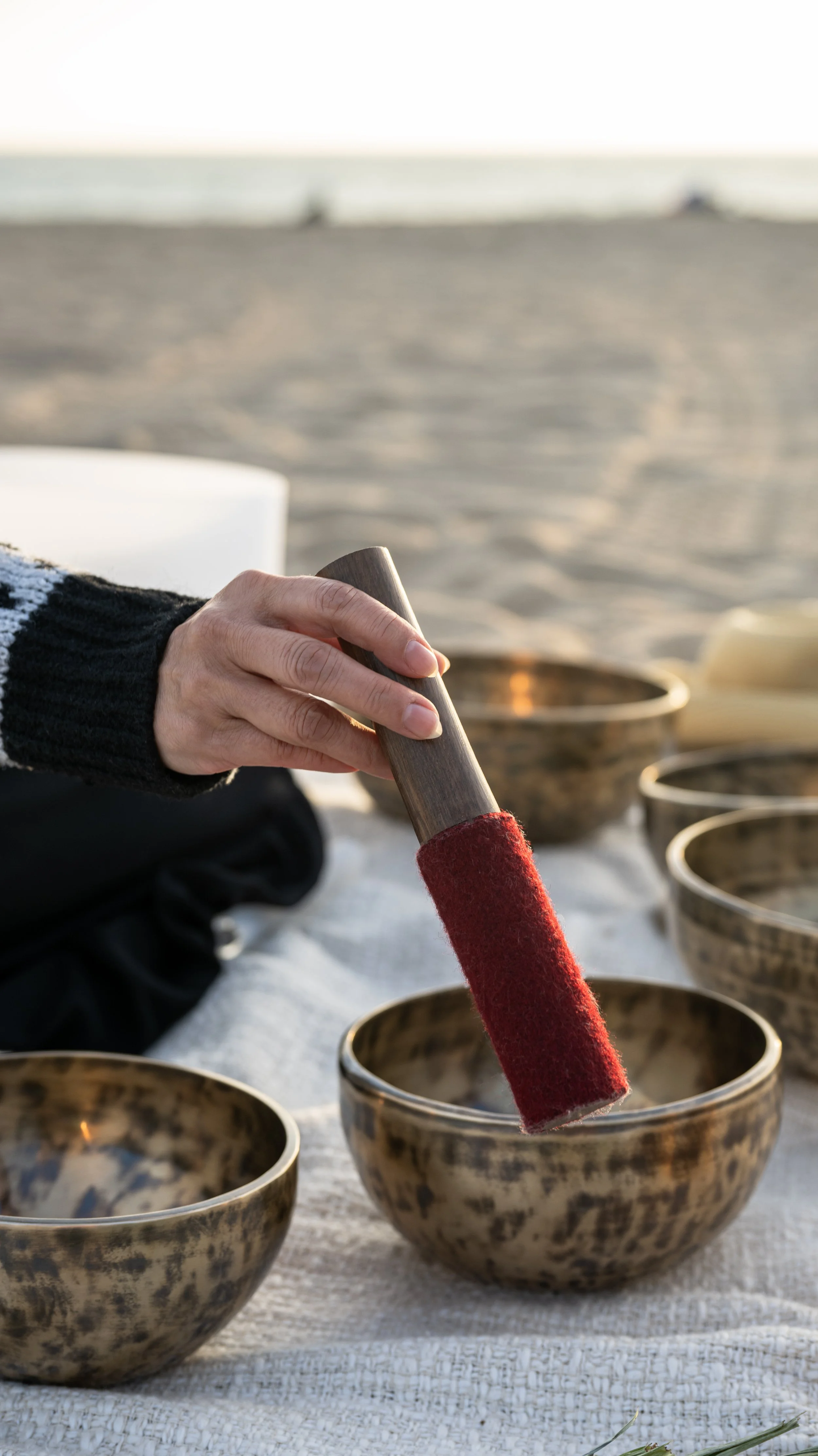 Close-up of a person's hand holding a mallet, striking singing bowls on a white cloth at the beach during sunset.