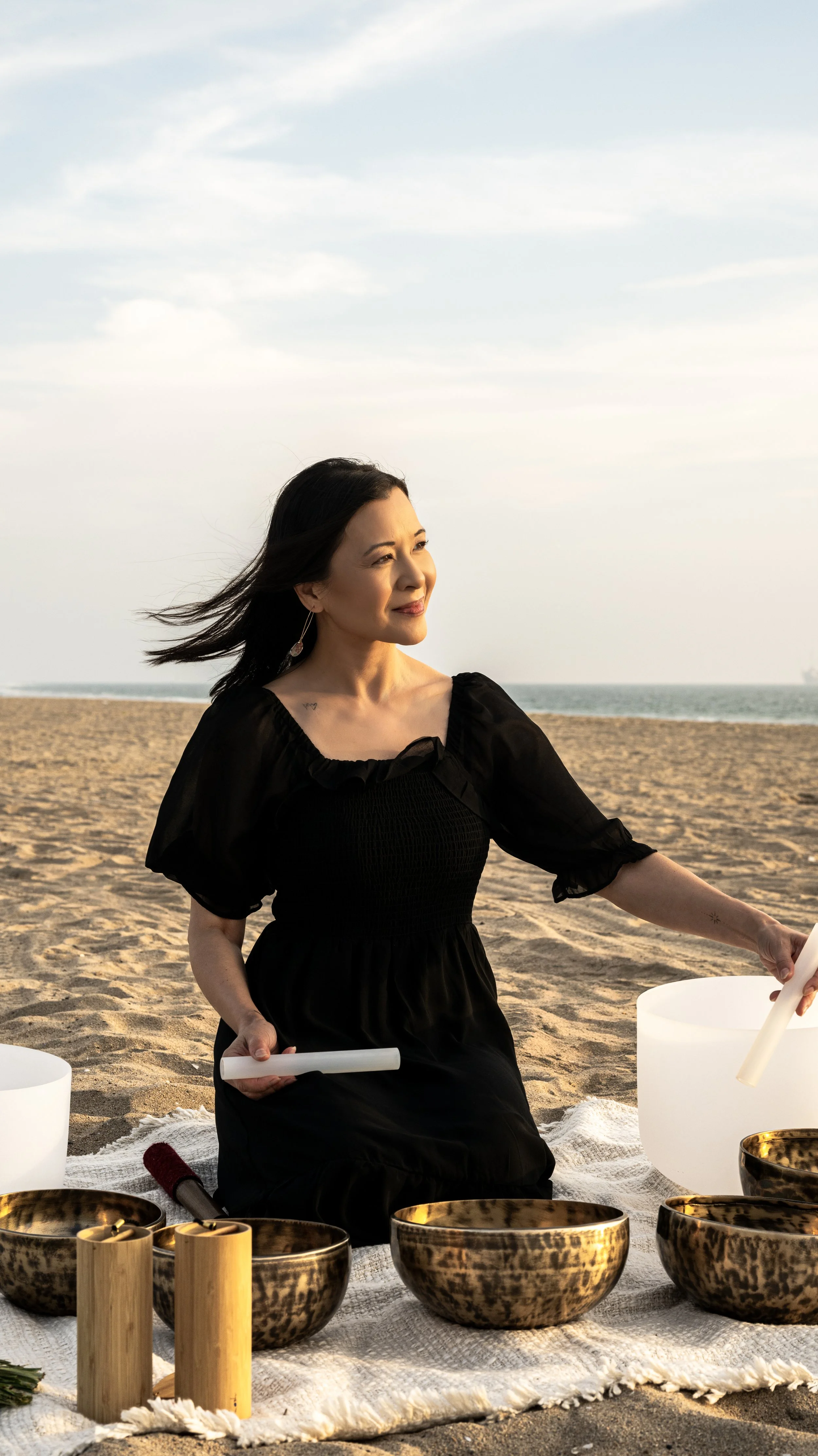 Woman in a black dress playing crystal singing bowls on the beach at sunset.