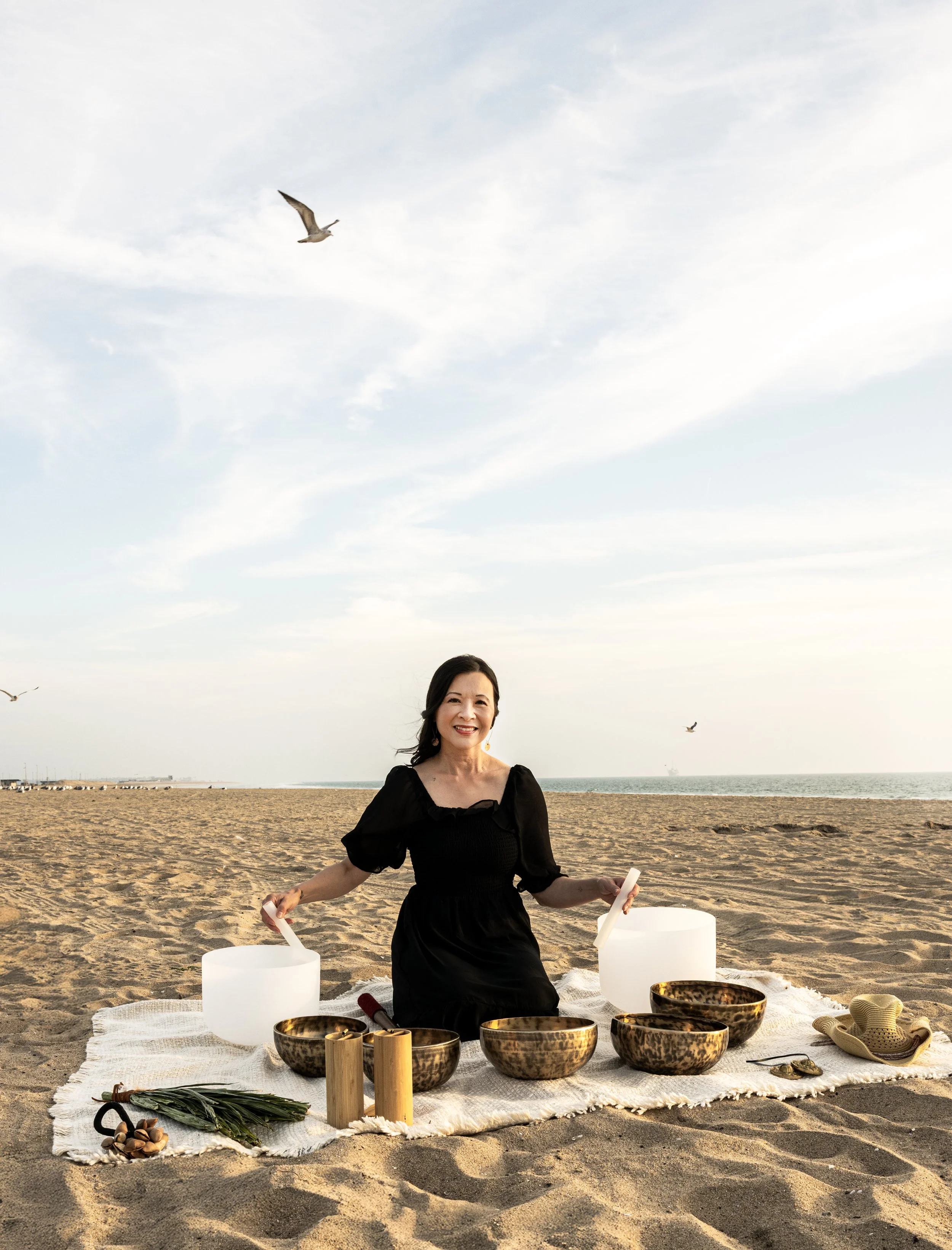 A woman in a black dress sitting on a white mat on a sandy beach, surrounded by singing bowls, candles, and other spiritual items, with seagulls flying in the sky and the ocean in the background.