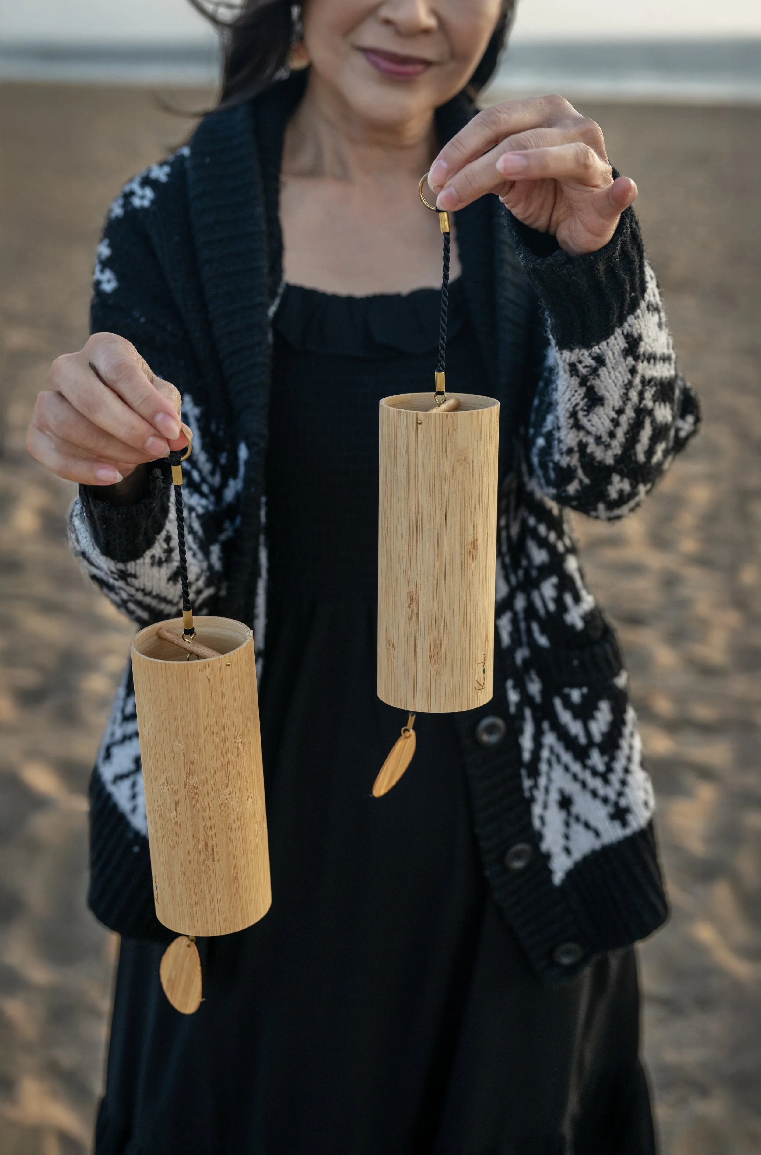 A woman holding two bamboo wind chimes with wooden clappers on a beach during sunset.