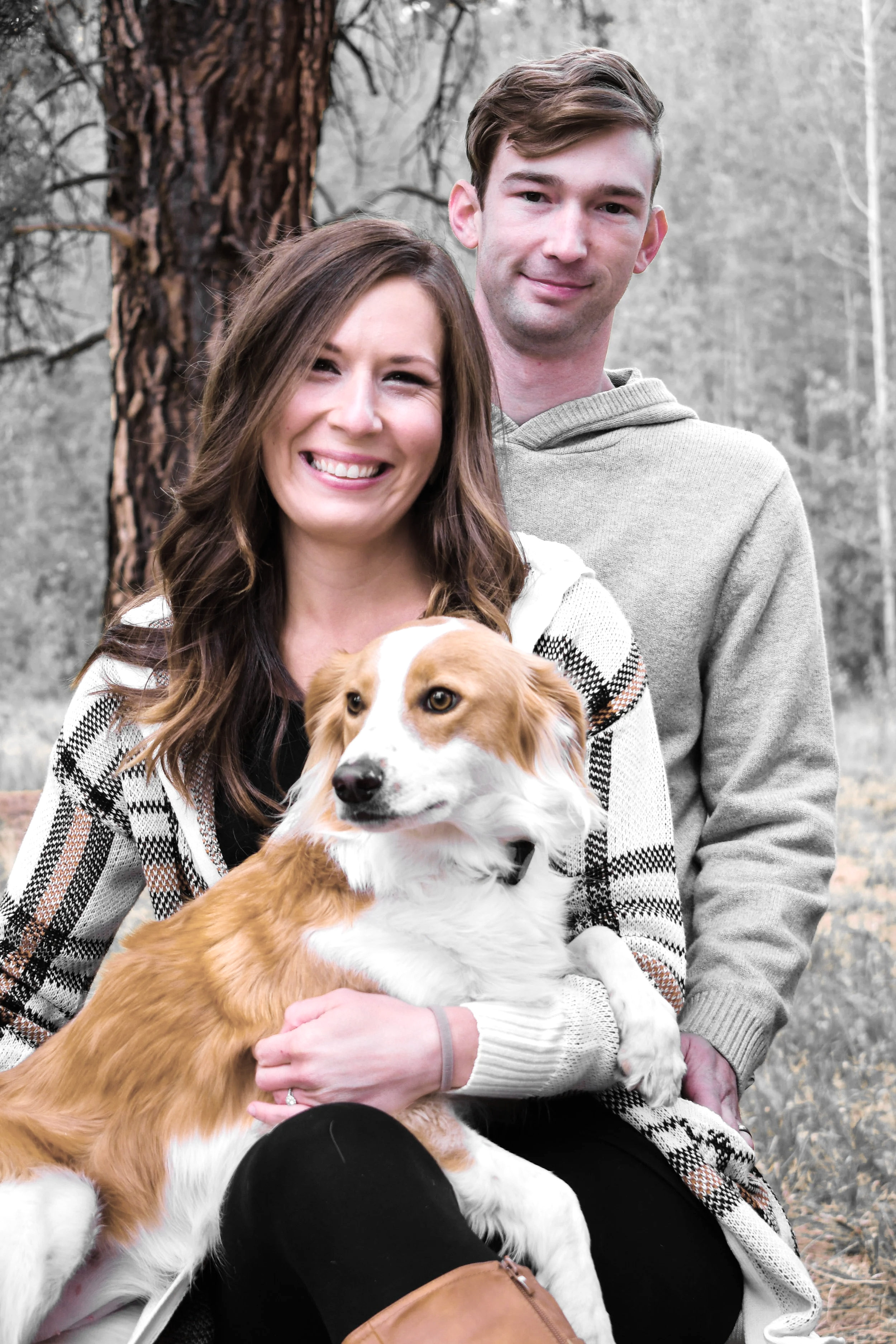 A smiling woman holds a golden retriever while sitting outdoors with a man in a gray hoodie in the background, near trees.