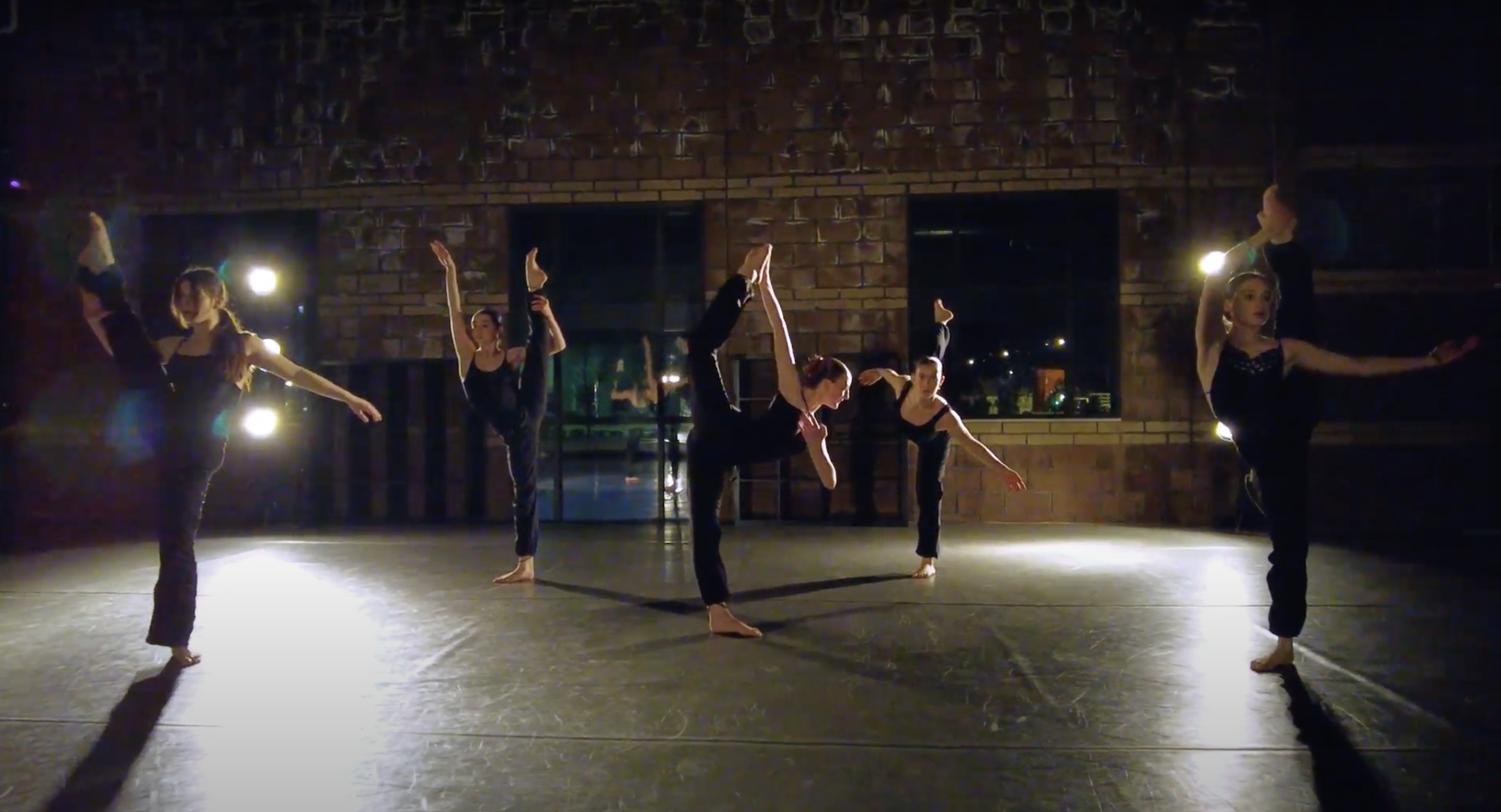 5 dancers dressed in black strike a finishing pose to their dance in a studio rehearsal. Each pose shows off their extension and flexibility. The studio is dark, but dramatically lit from behind from two exposed boom lights.