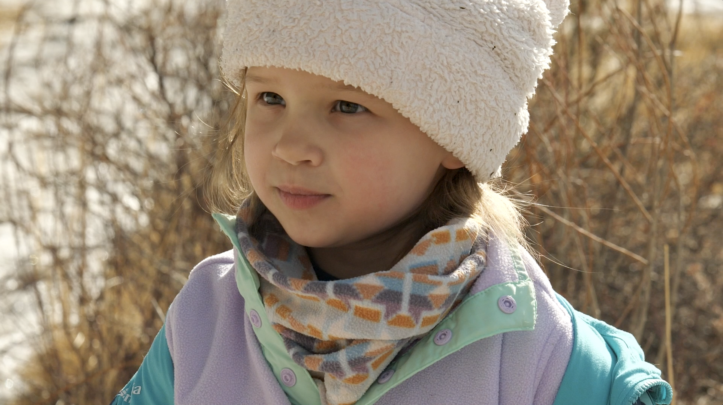 A young girl in a white hat, colorful scarf, and purple jacket looks off into the distance. In the background is a snowy, grassy trail.