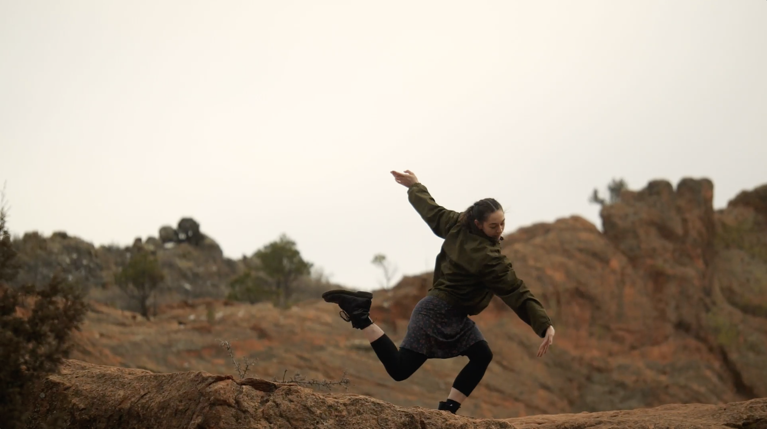 a dancer moves through an angular pose as if they are running in slow motion. she wears a green jacket, blue skirt, black tights, and black boots. she is on top of a red rock and surrounded by red rocks.