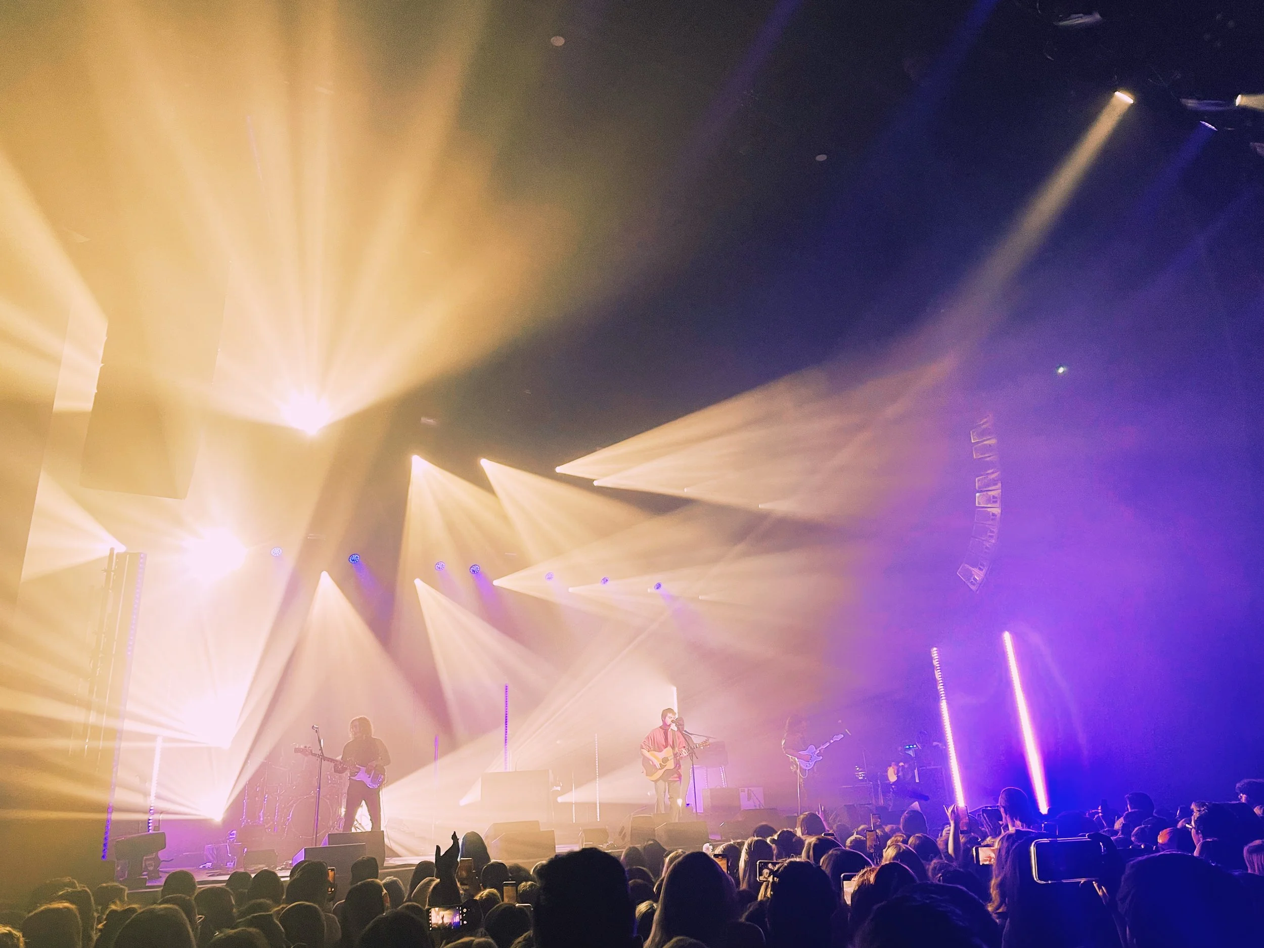 A band onstage with guitars sings to the crowd while golden beams of light stretch out from behind them.