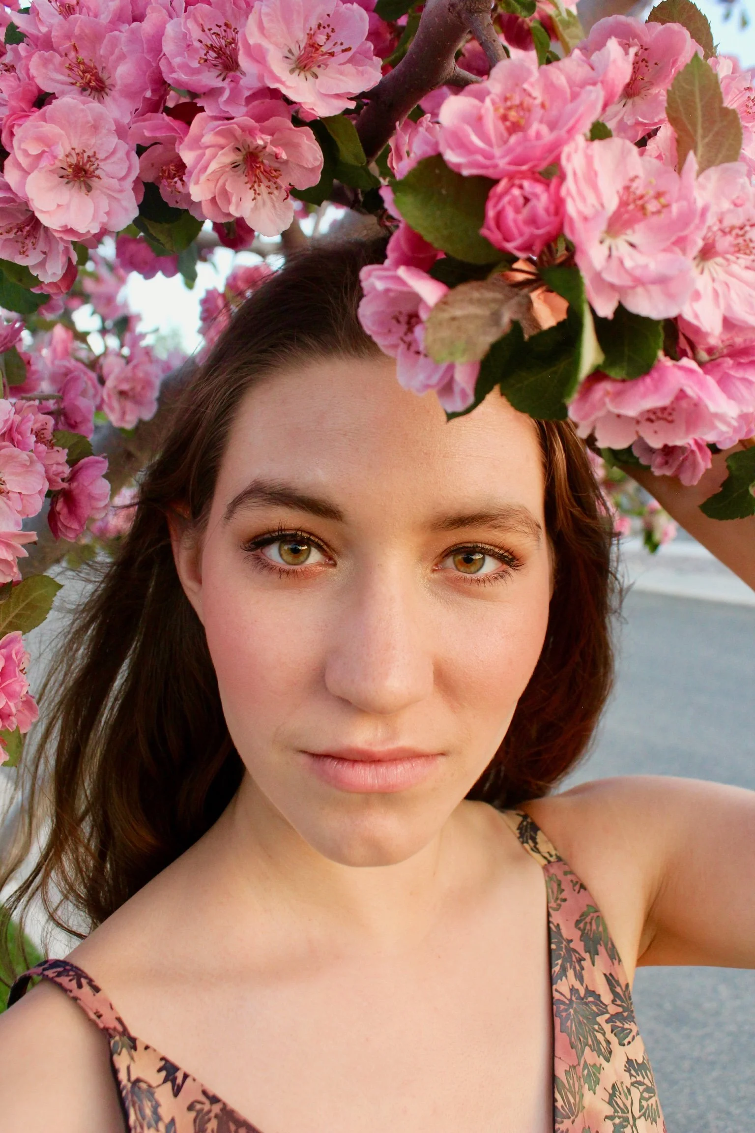 A headshot of a young woman with long brown hair and hazel eyes stands beneath a tree of pink blossoms