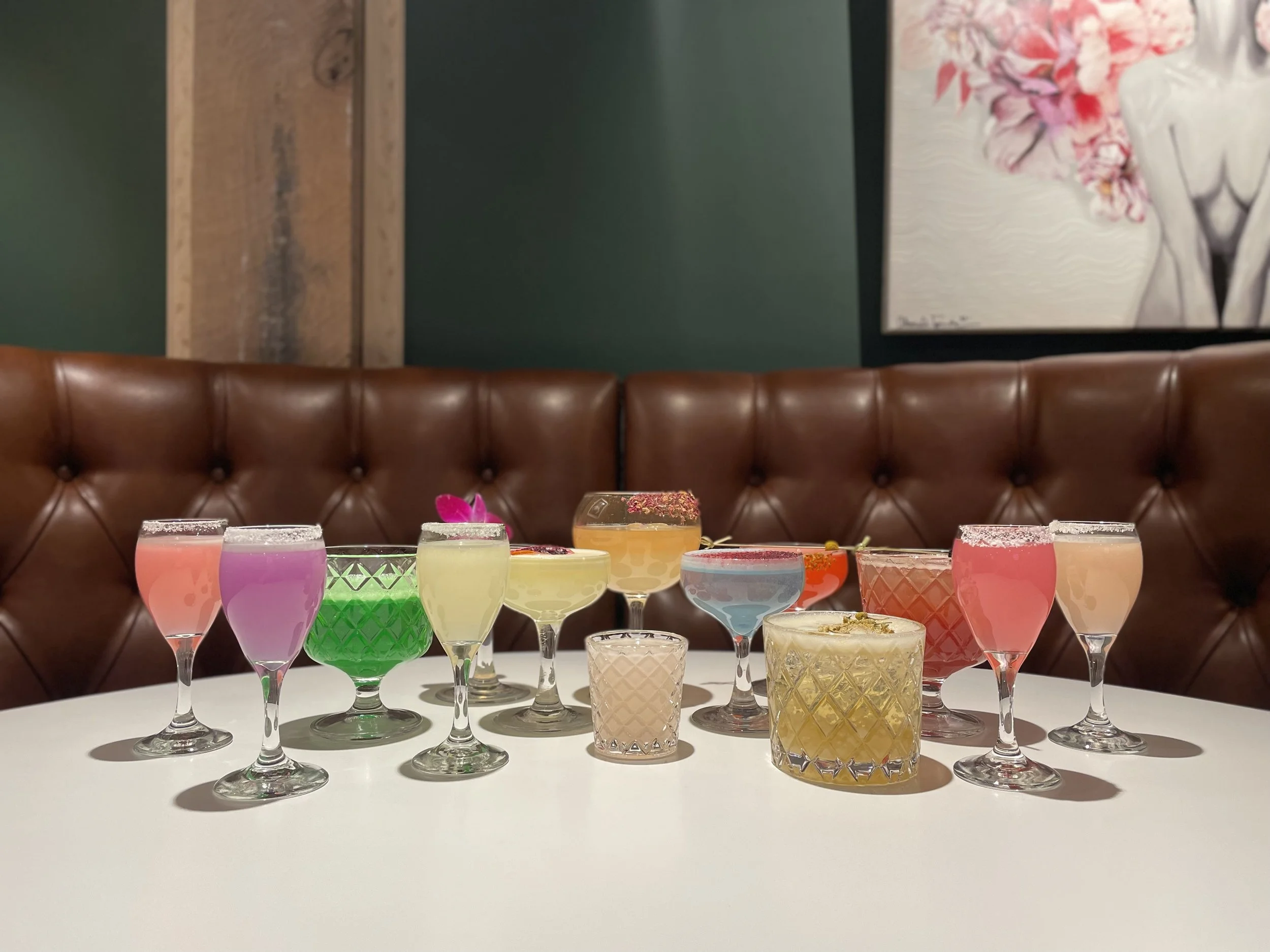 an assortment of unique, colorful cocktails atop a white table with brown leather booth seating.