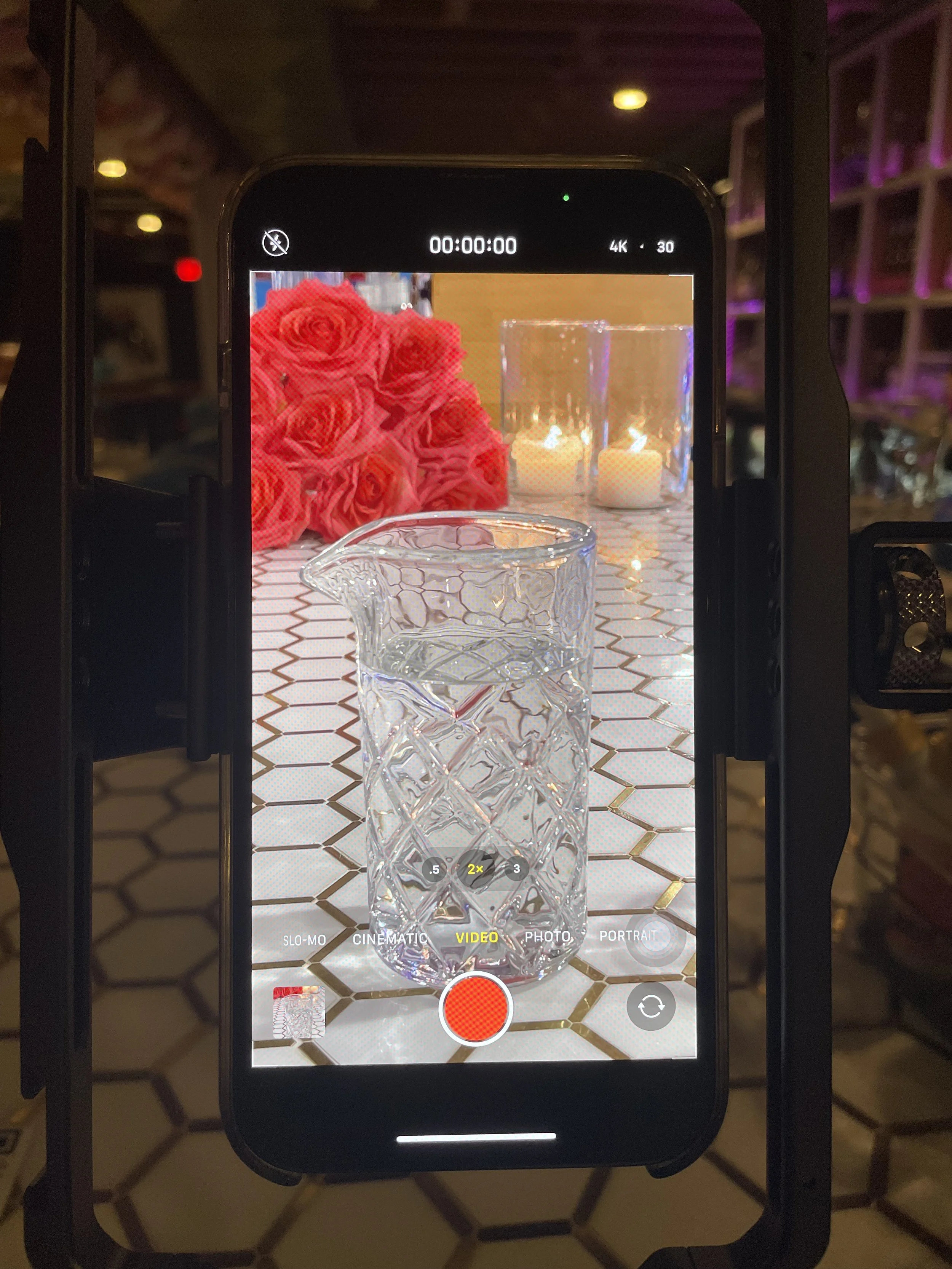 photo of a recording iPhone on a tripod about to capture a video of a glass on a white tile bar top with pink roses and white candle sin the background