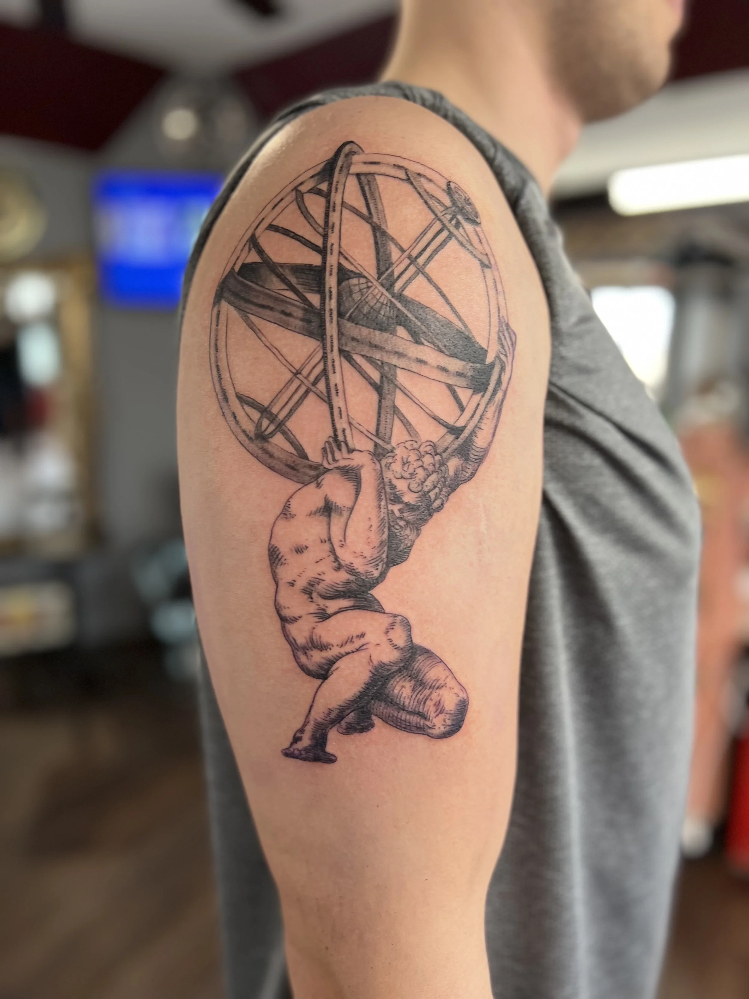 Tattoo of a muscular man holding up a large armillary sphere with a small airplane inside it, on a person's upper arm.
