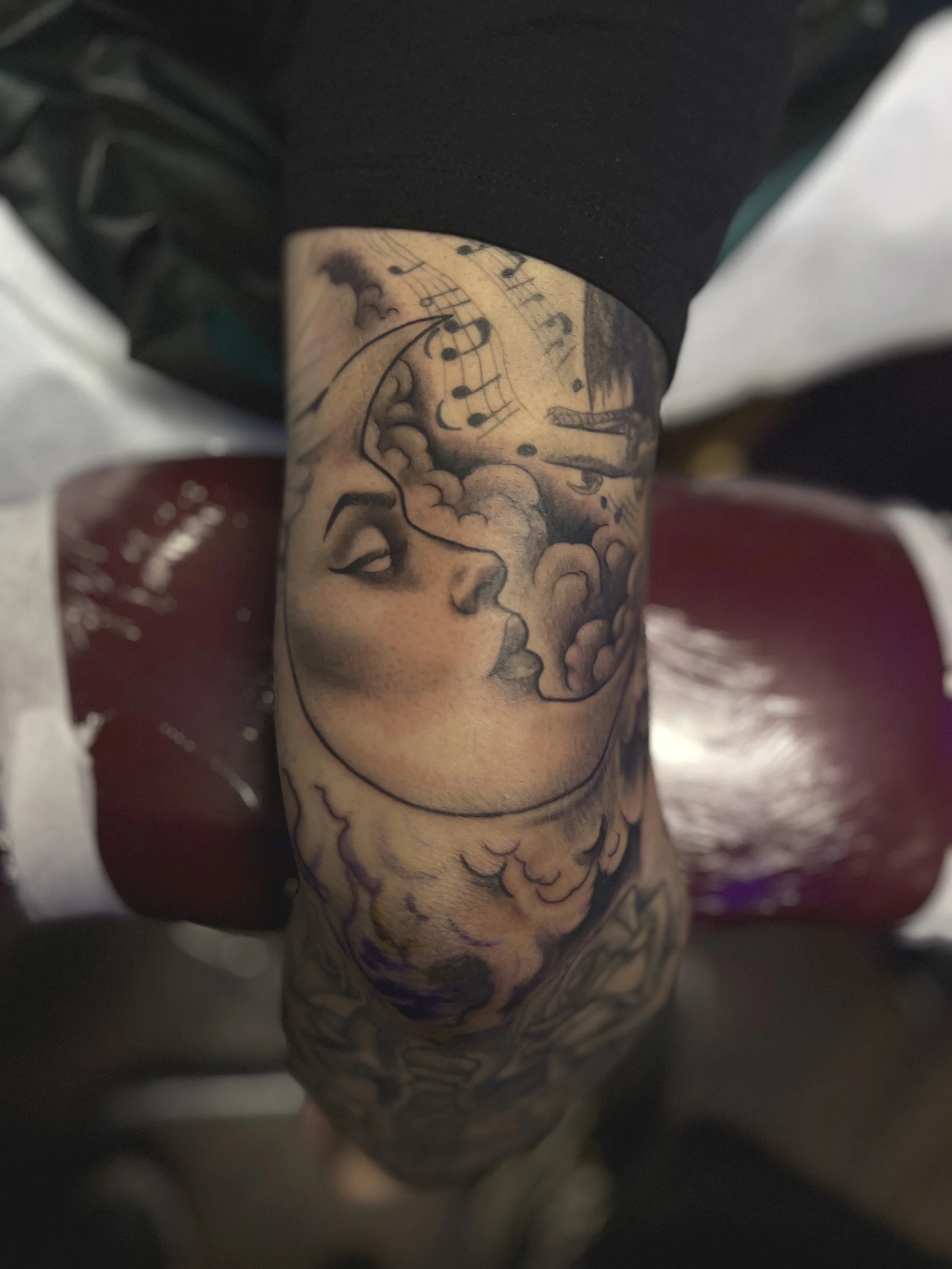 Tattoo of a woman's face with closed eyes, a moon with clouds, and musical notes on the forearm.