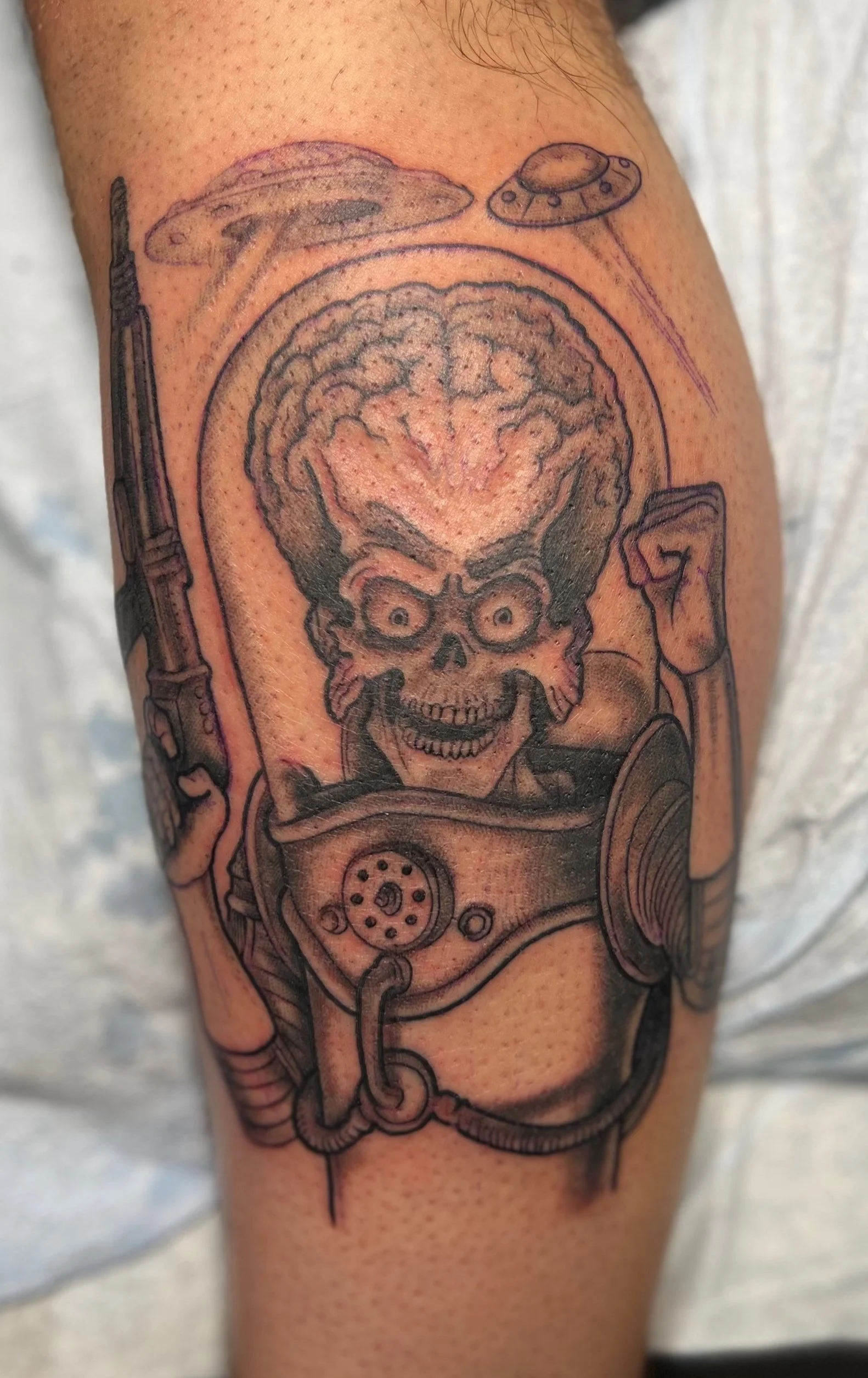 Tattoo of a skeleton with an exposed brain, wearing a space suit, holding a gun, with UFOs in the background.
