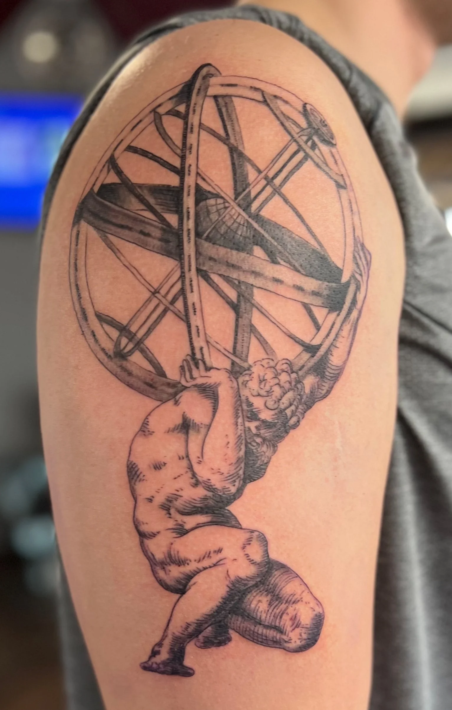 Tattoo of a muscular man holding up a large armillary sphere with a small airplane inside it, on a person's upper arm.