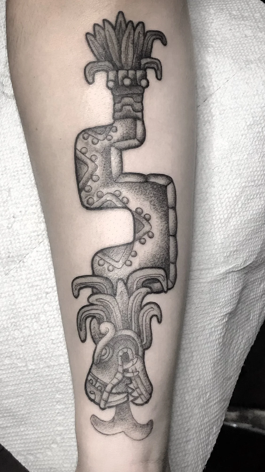 A tattoo of a stylized, elongated figure with a face, flowing hair, and intricate patterns, resembling a totem or mythical creature, on a person's forearm. Flying serpent.