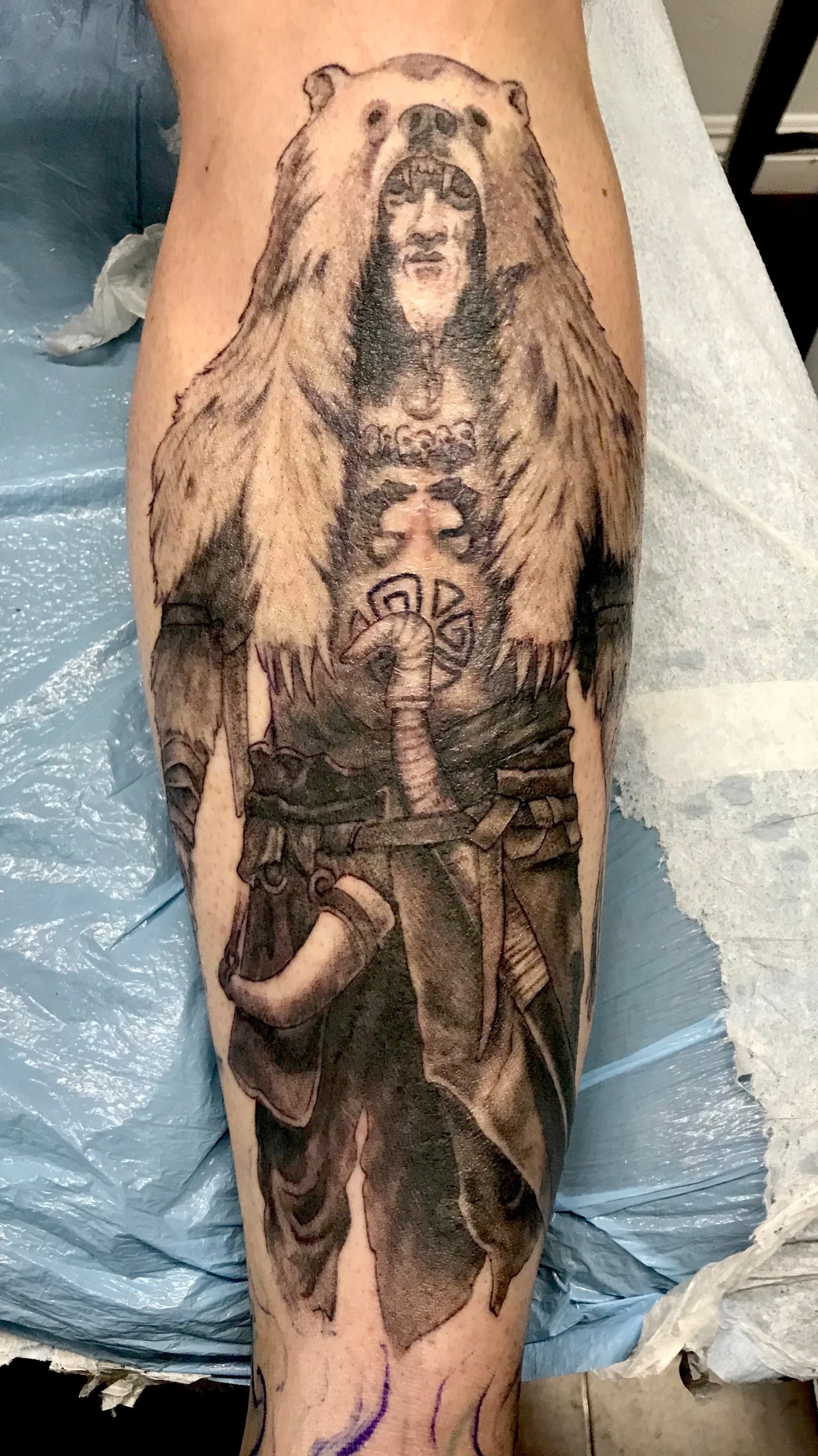 Black and gray tattoo of a Native American warrior dressed in traditional clothing, with a headdress and a snake wrapped around his chest, on a person's leg.