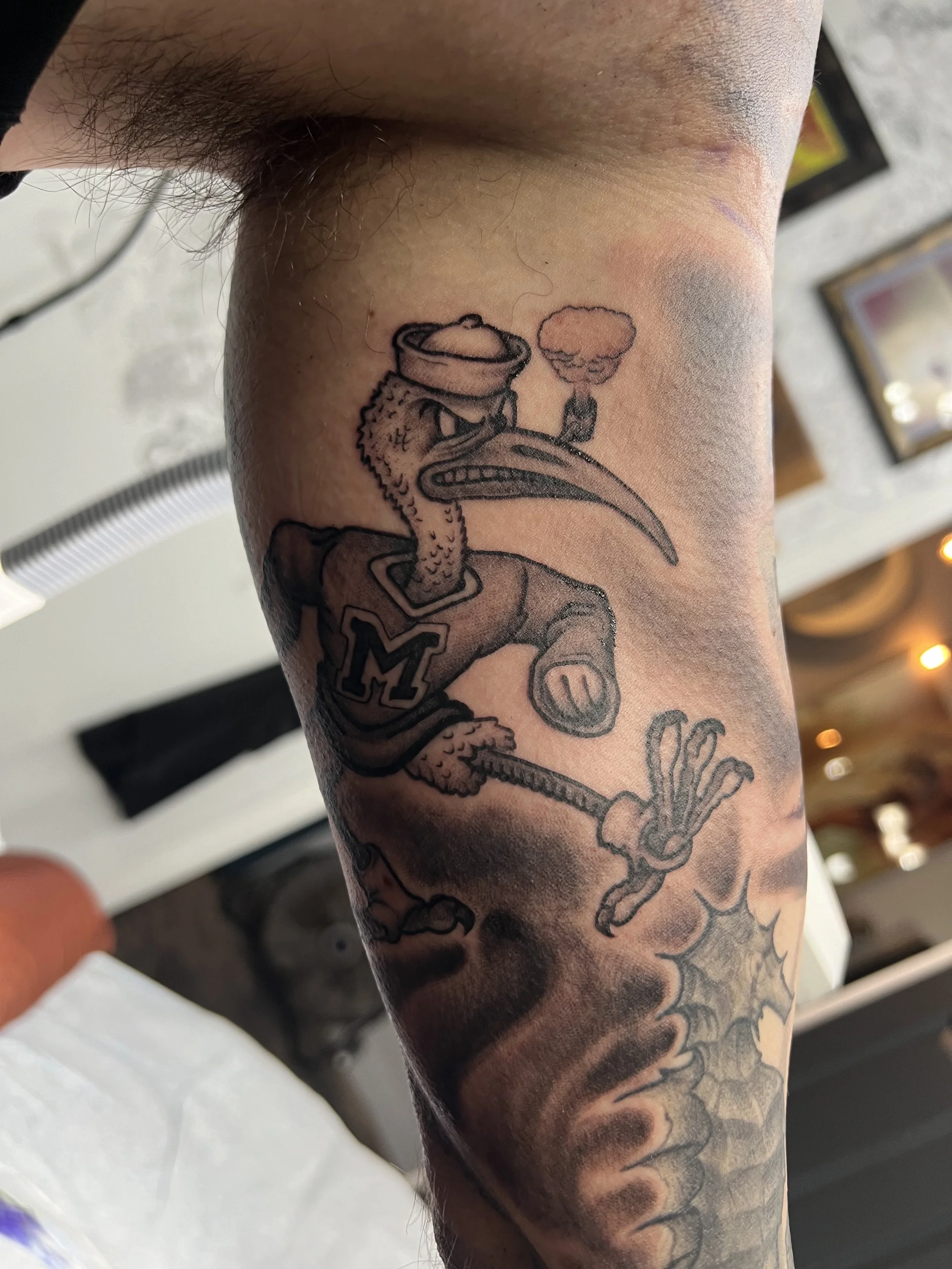 Tattoo of a cartoon bird dressed as a student or athlete, holding a basketball, with steam rising from its beak, wearing a jacket with the letter 'M', and surrounded by clouds and a spider web design.