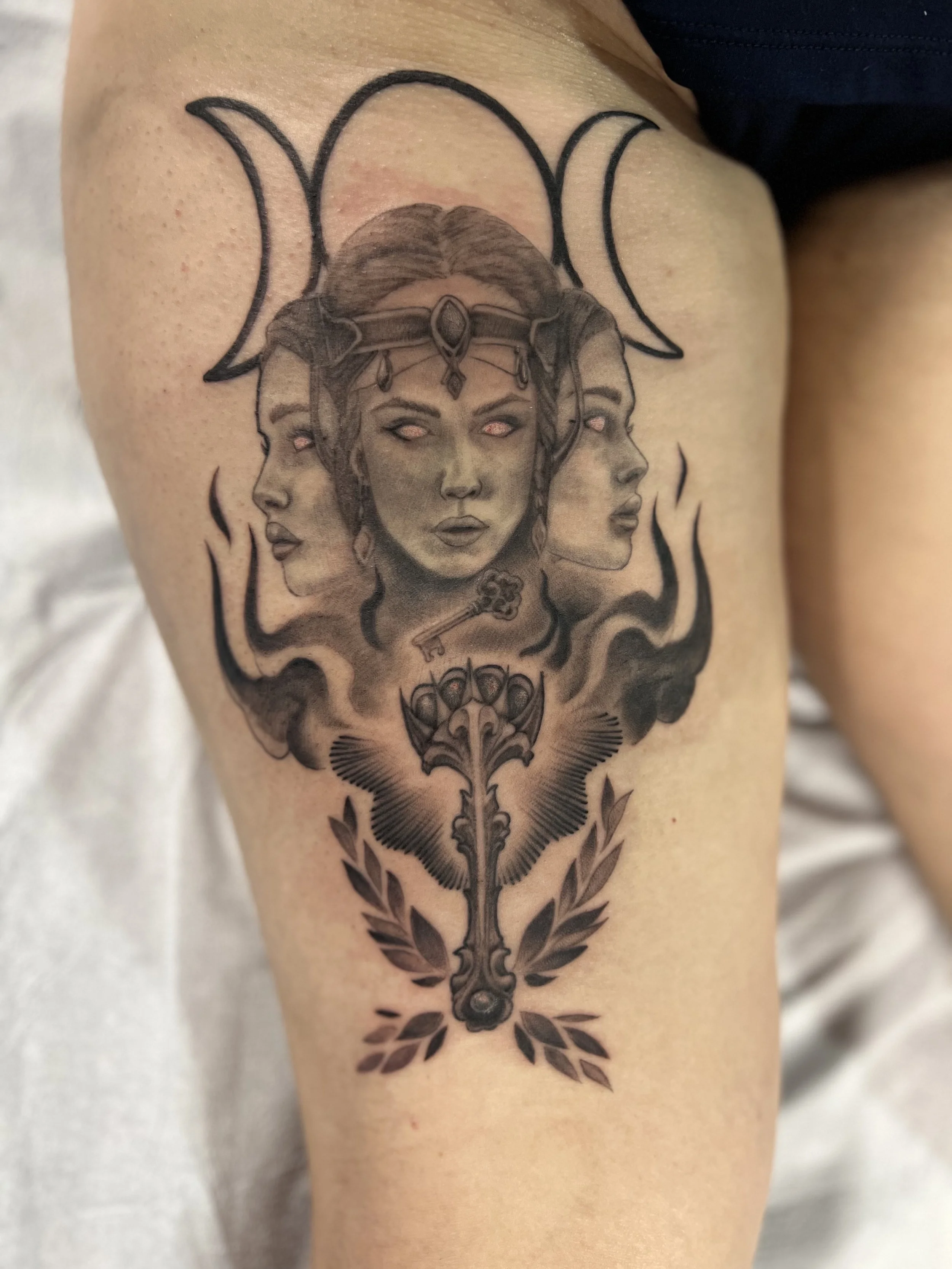 Tattoo artwork featuring three female faces with closed eyes and headbands, a moon phase symbol above, a key hanging from the central face, a decorative spear with wings and laurel leaves below.