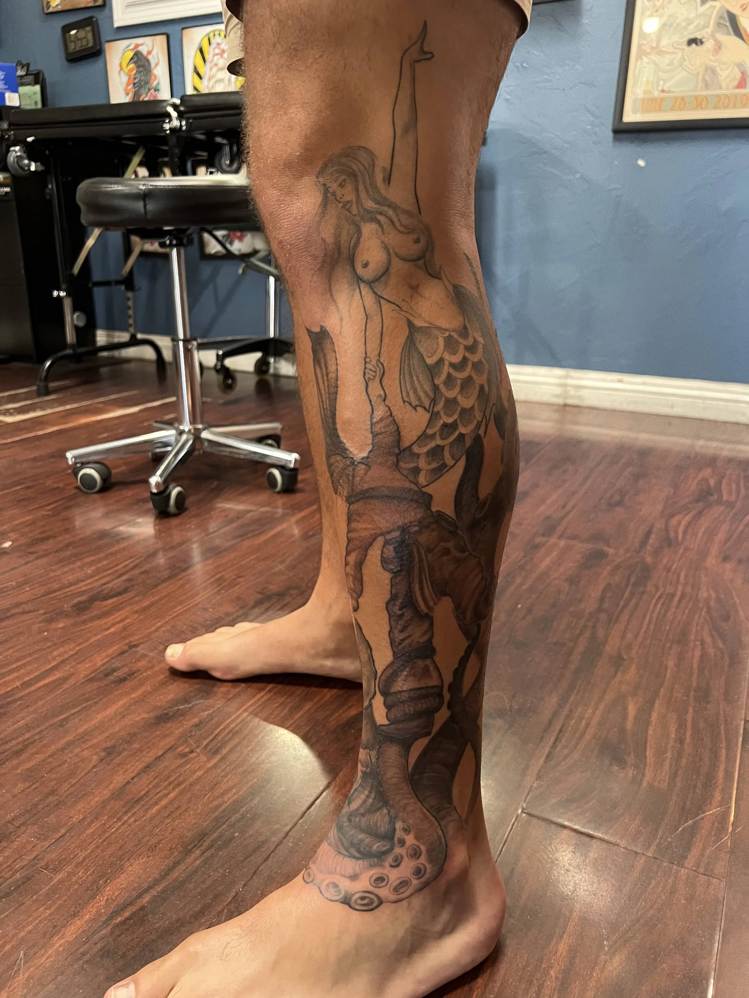 Tattoo of a mermaid with a whale tail on a person's leg, featuring a woman with long hair holding a fishing hook.