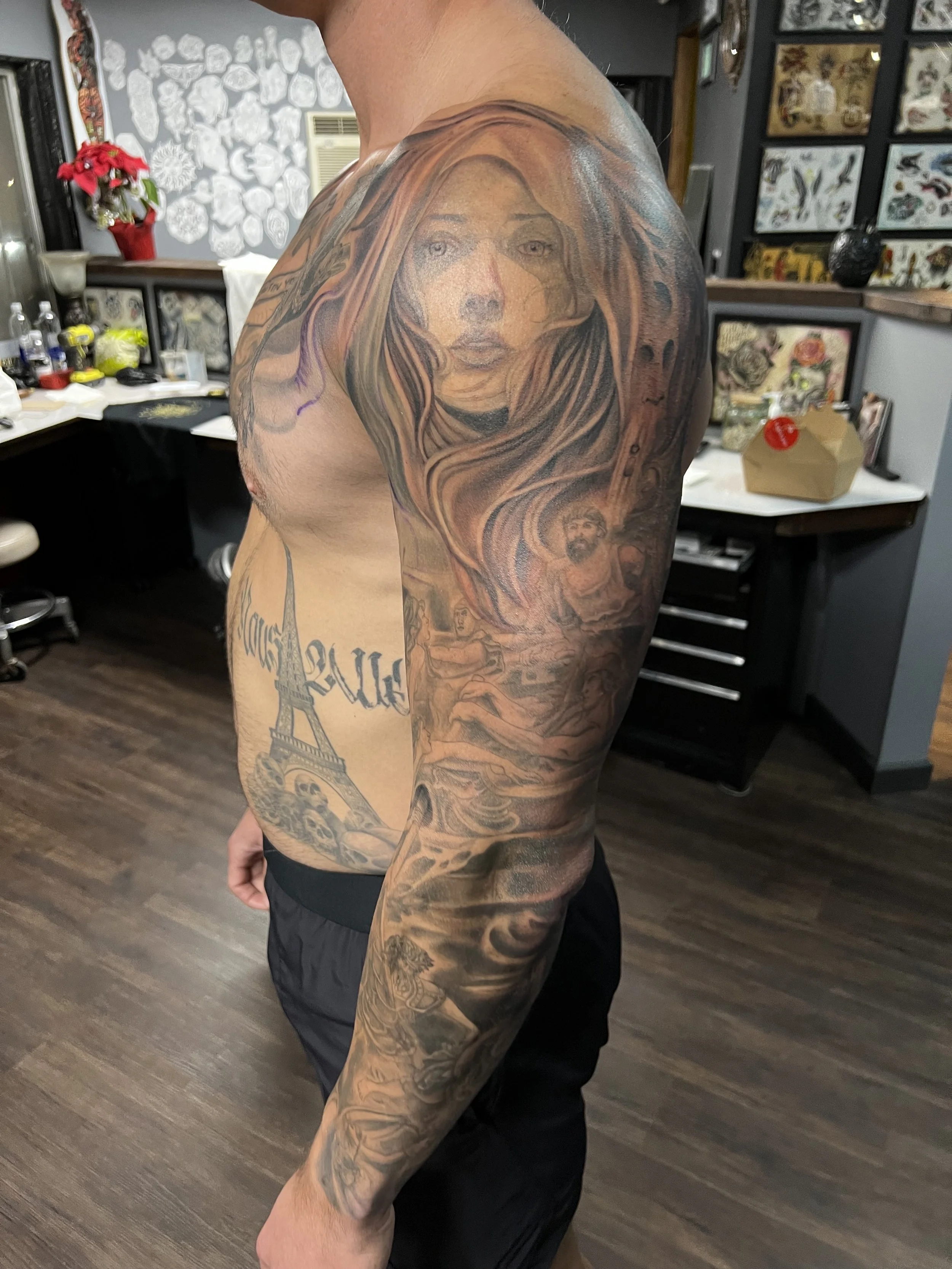 A man with a large, detailed tattoo of a woman's face with flowing hair covering his upper chest, shoulder, and arm. The tattoo features realistic shading and intricate line work.