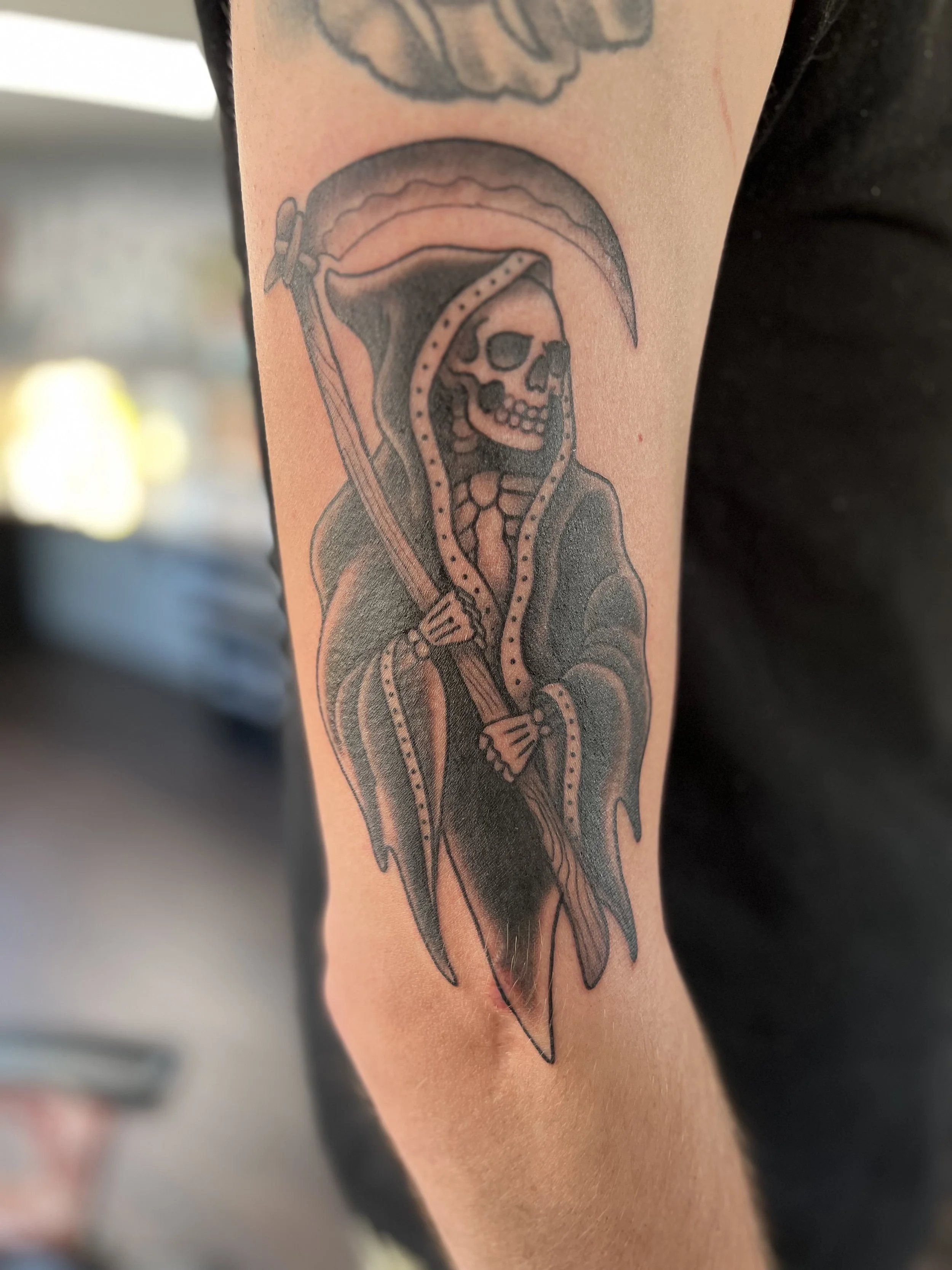Tattoo of a Grim Reaper with a skull face, wielding a scythe, wearing a hooded cloak.