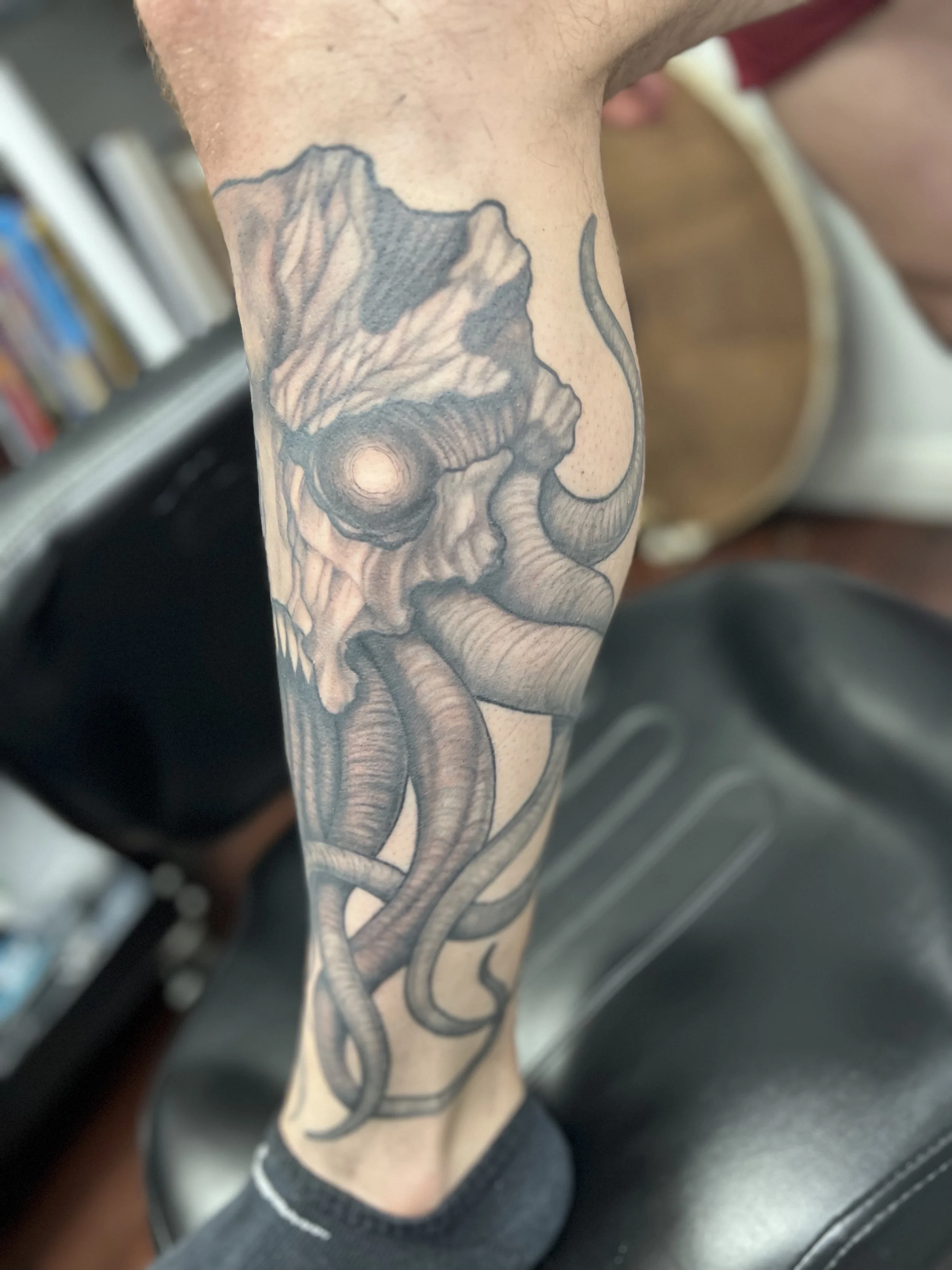 Detailed black and gray tattoo of a skull with angry eyes and snake-like tentacles on a person's lower leg.