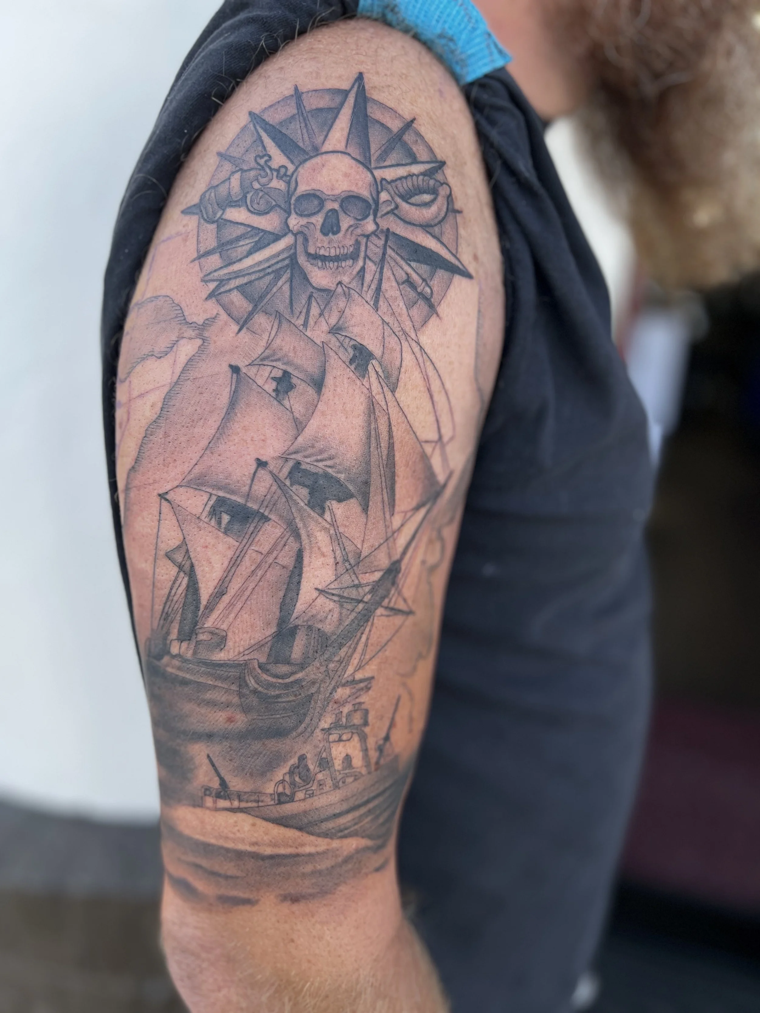 Tattoo of a sailing ship, a skull with a sunburst background, and clouds on a person's upper arm.